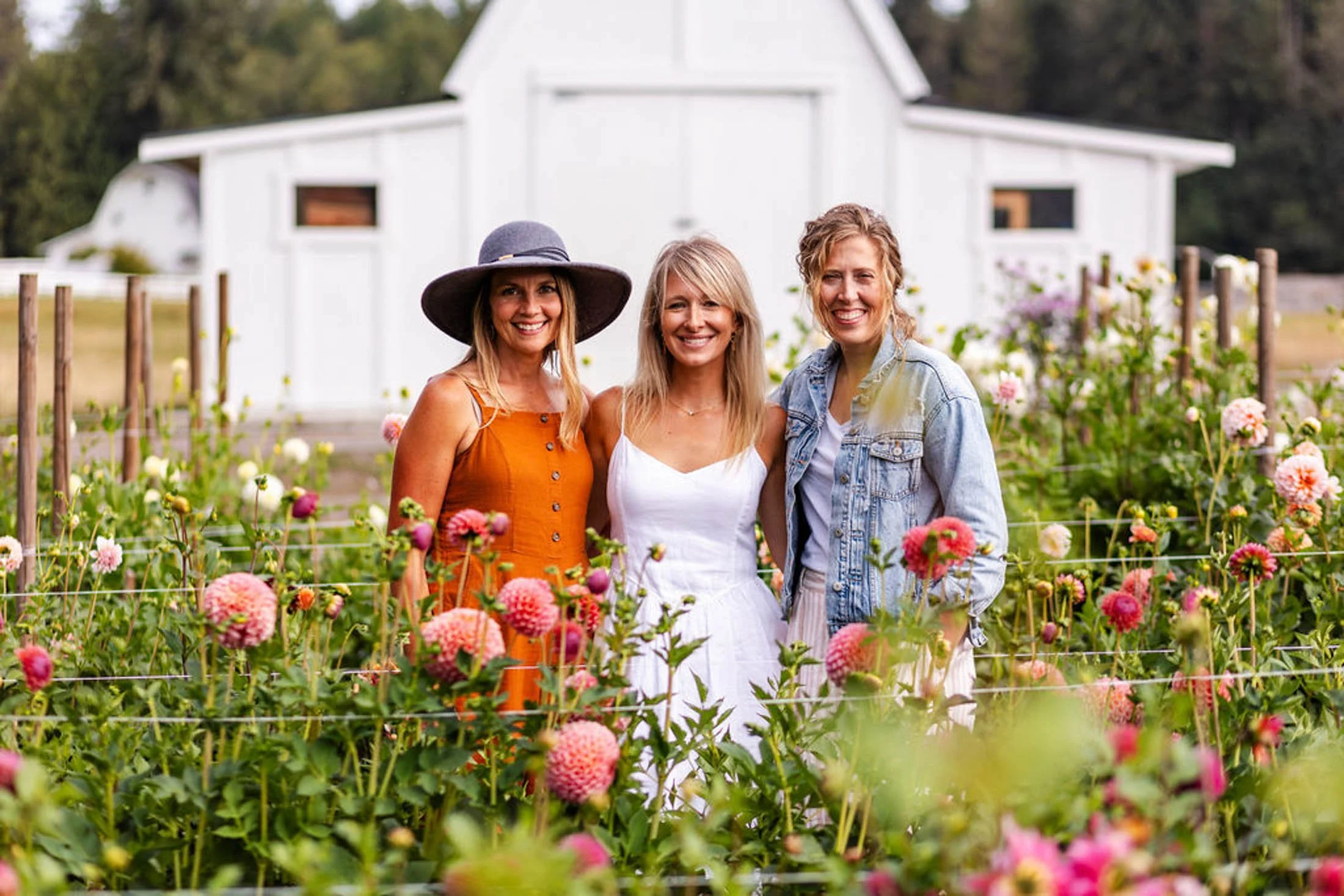 Three women standing in a flower garden with a white barn in the background, smiling and enjoying the outdoors.
