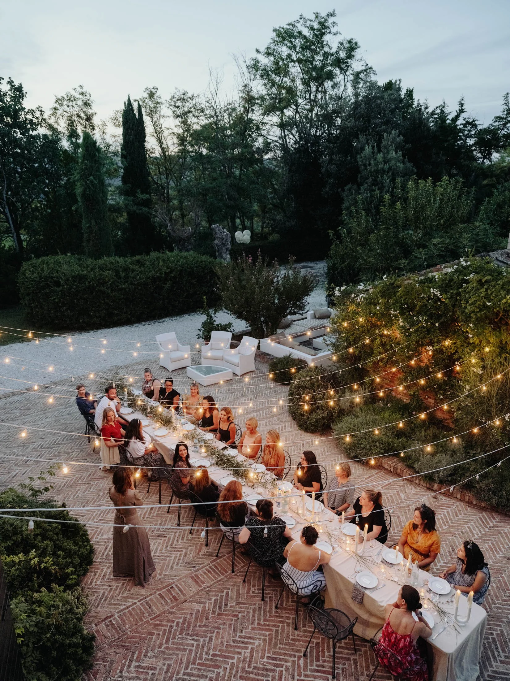A large outdoor dinner party with many attendees seated at a long table decorated with candles and greenery, set on a brick patio surrounded by lush green trees and string lights at dusk.