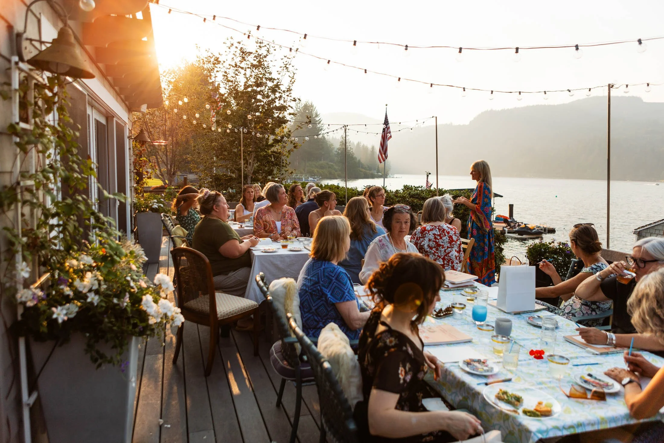 A group of people gathered around a long outdoor dining table by a lake at sunset, with string lights overhead and an American flag in the background.