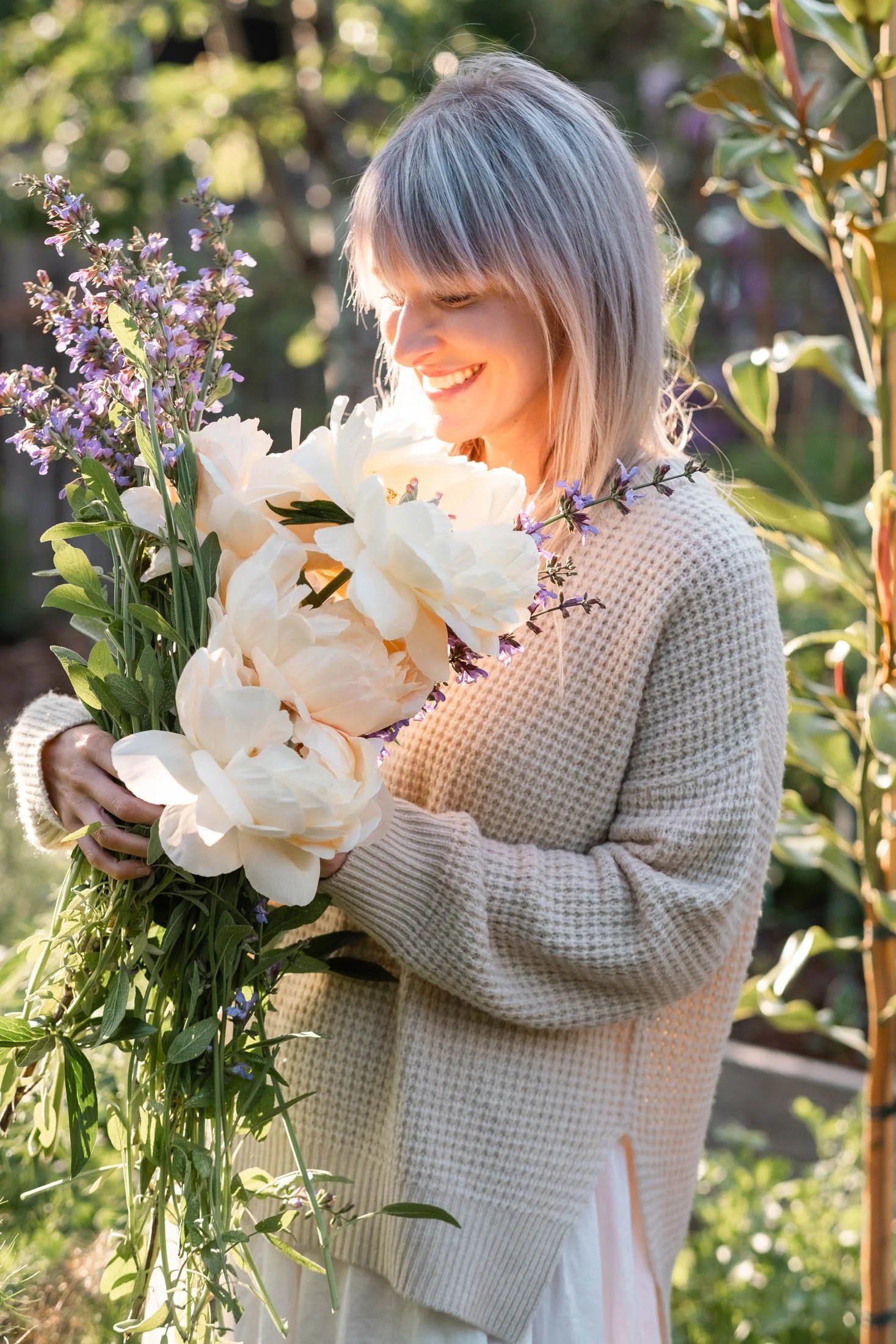 A woman is holding a bouquet of white and purple flowers in a garden with green foliage and sunlight.
