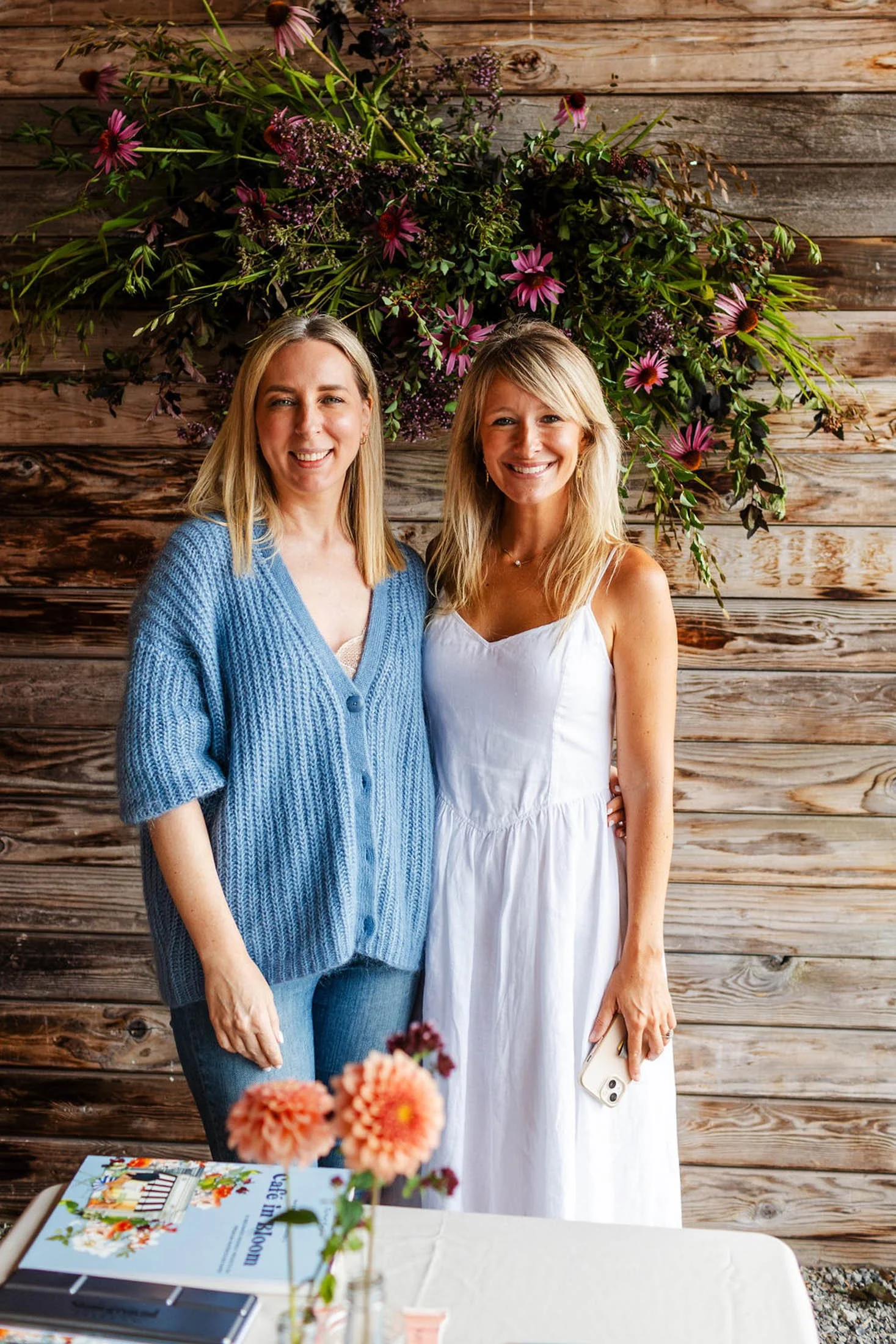 Two women smiling and posing in front of a wooden wall decorated with pink and purple flowers, with a table in the foreground that has a book, a vase of flowers, and some smartphones.
