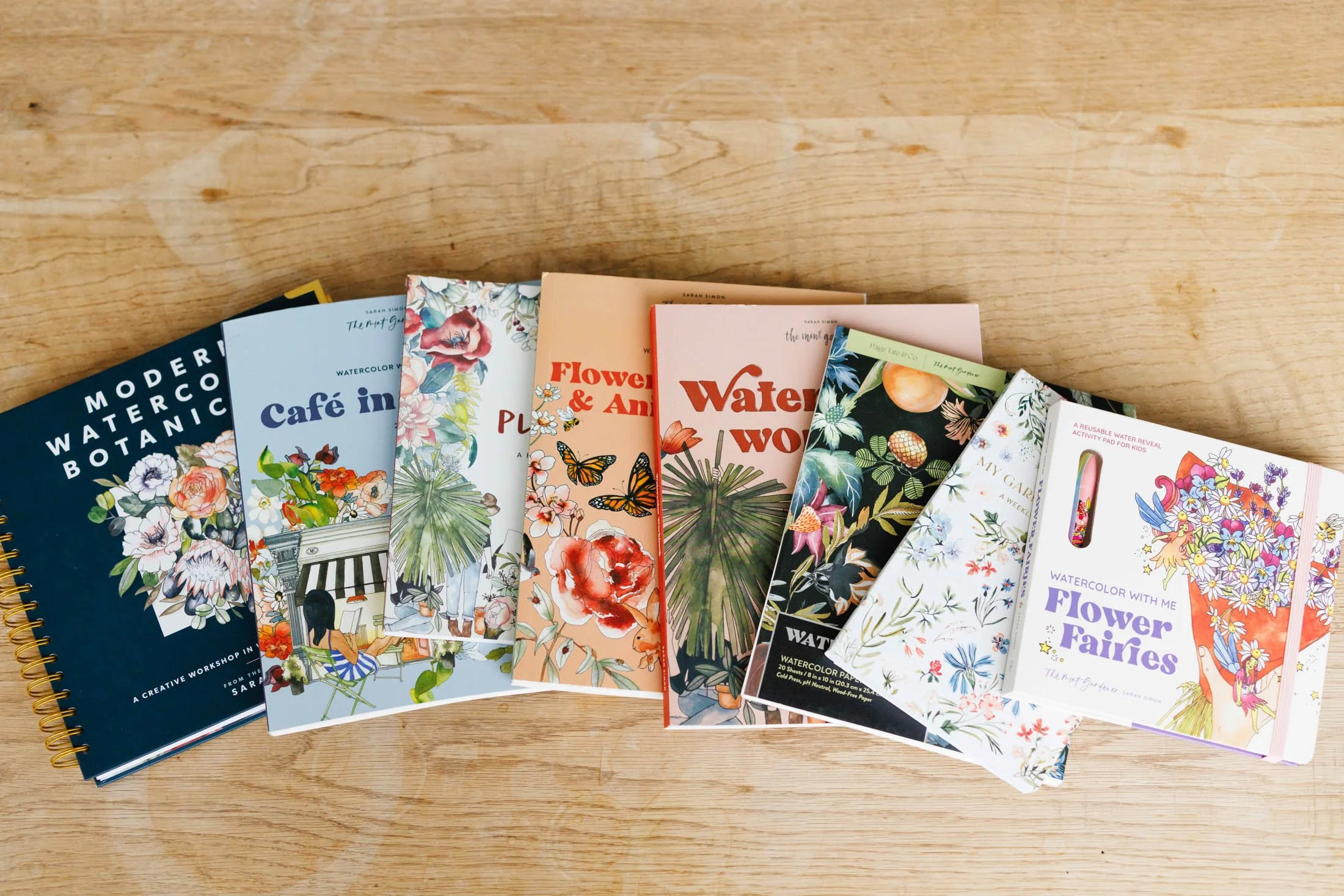A collection of watercolor and floral-themed books and activity pads spread out on a wooden surface.