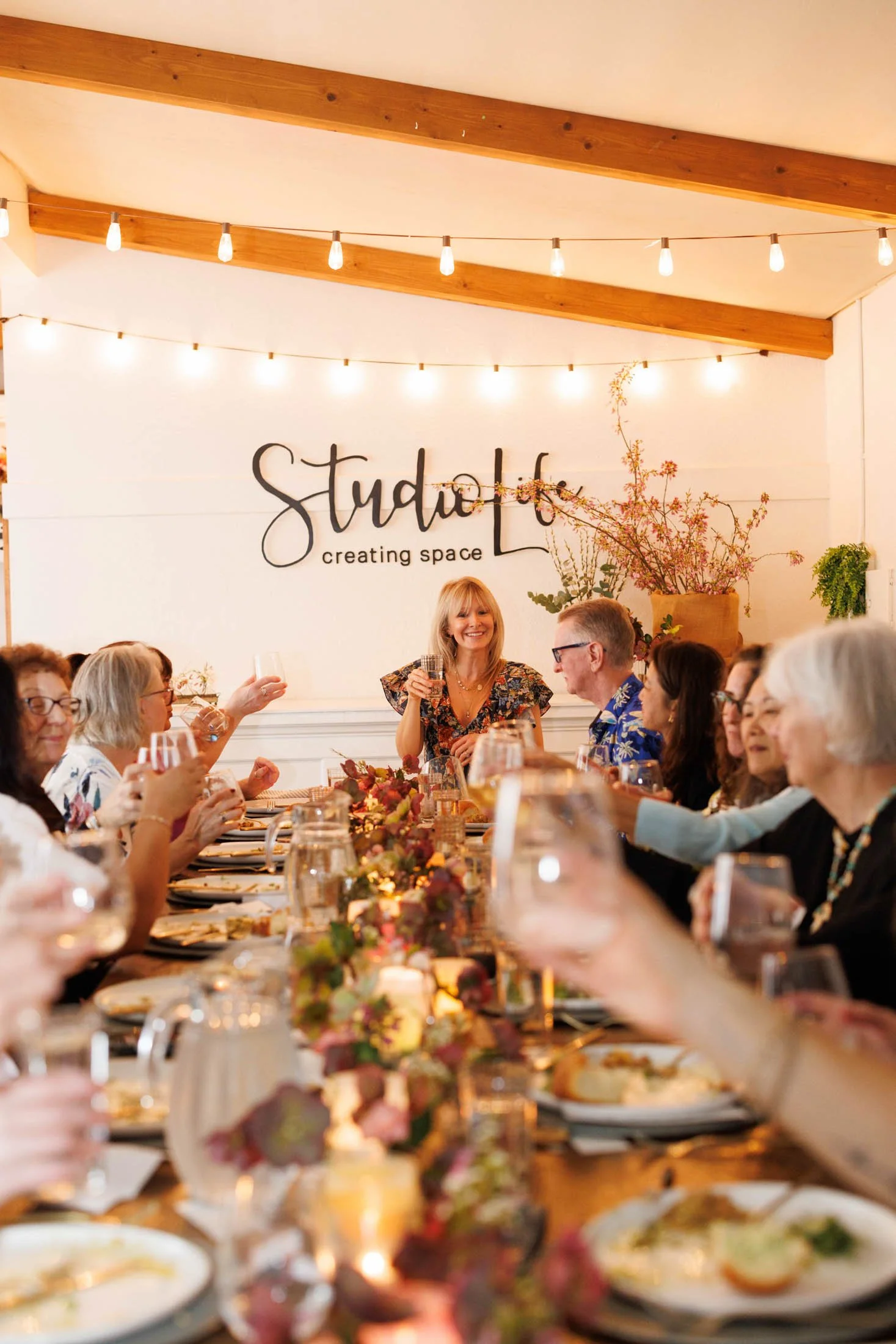 People gathered at a long dining table with plates, glasses, and floral centerpieces. The background features a wall with a sign that says 'studioL creating space' and string lights. The setting appears warm and festive.