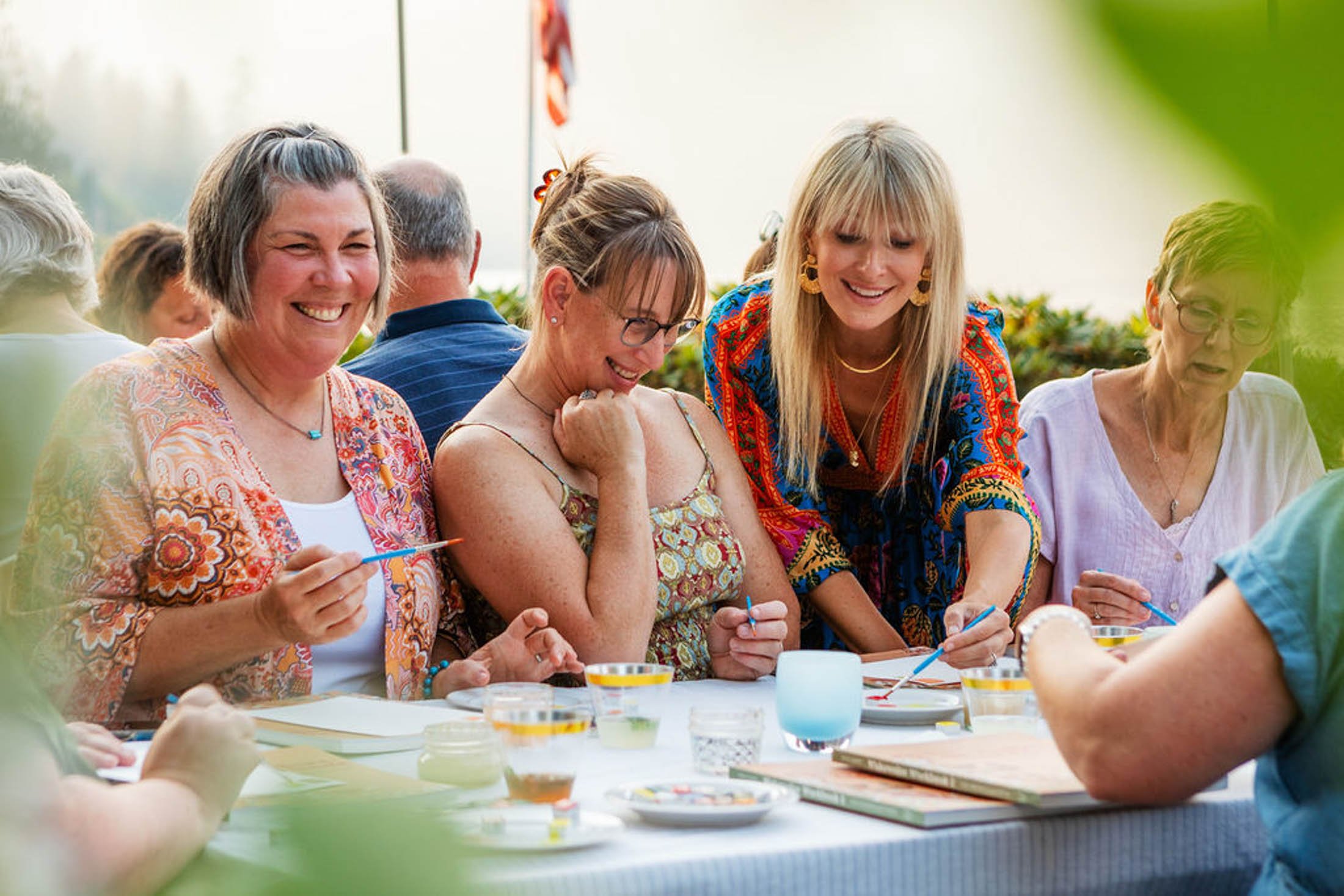 Group of women smiling and engaging in an art activity outdoors at a table with paints and brushes.