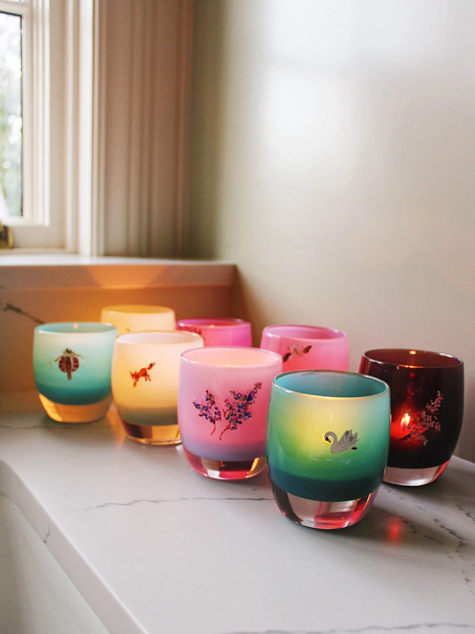 Colored glass candle holders with animal and nature designs arranged on a white windowsill in a bright room.