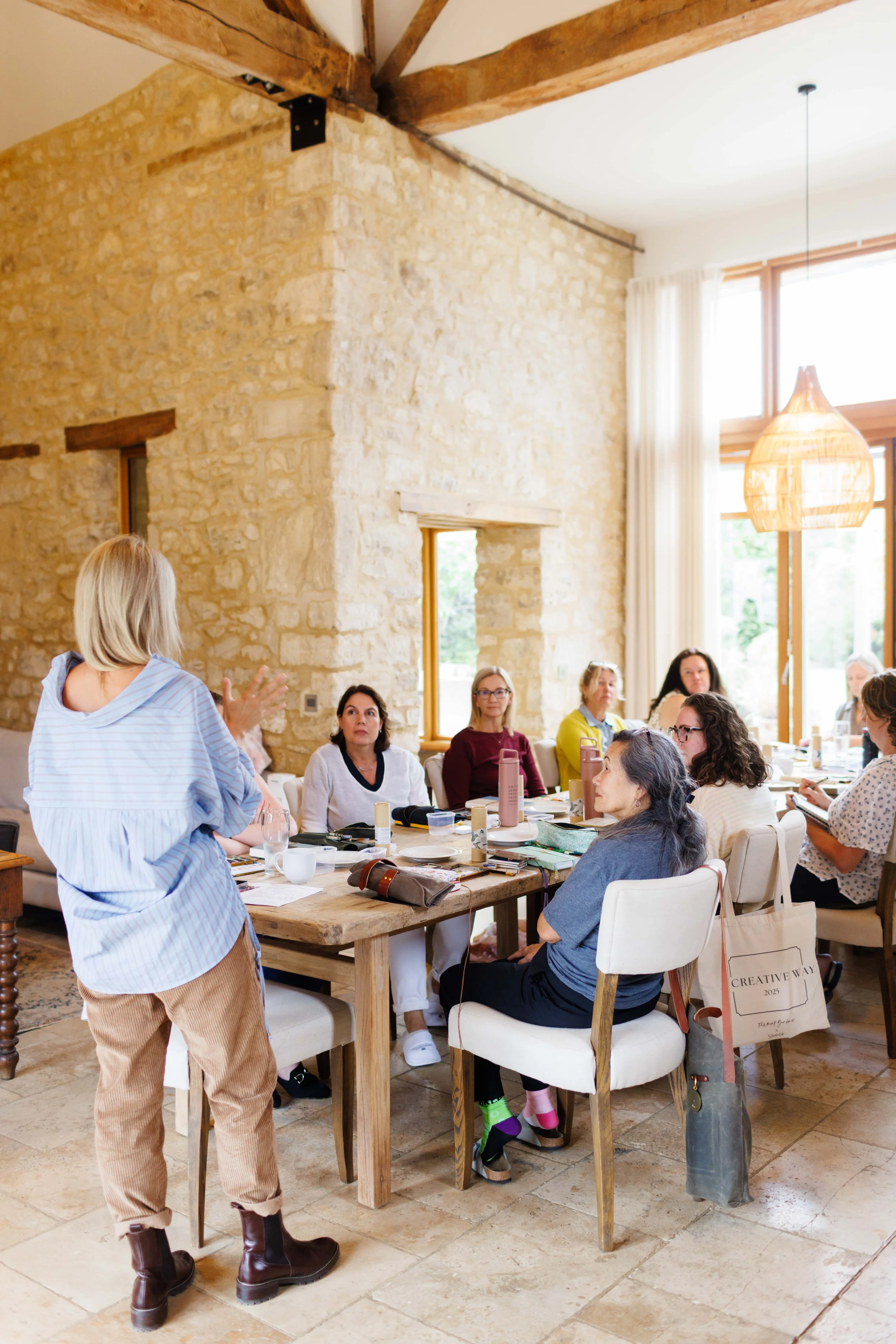 A woman with short blonde hair and a striped shirt is standing at a table, speaking to a group of women seated around the table, in a bright room with stone walls and large windows.