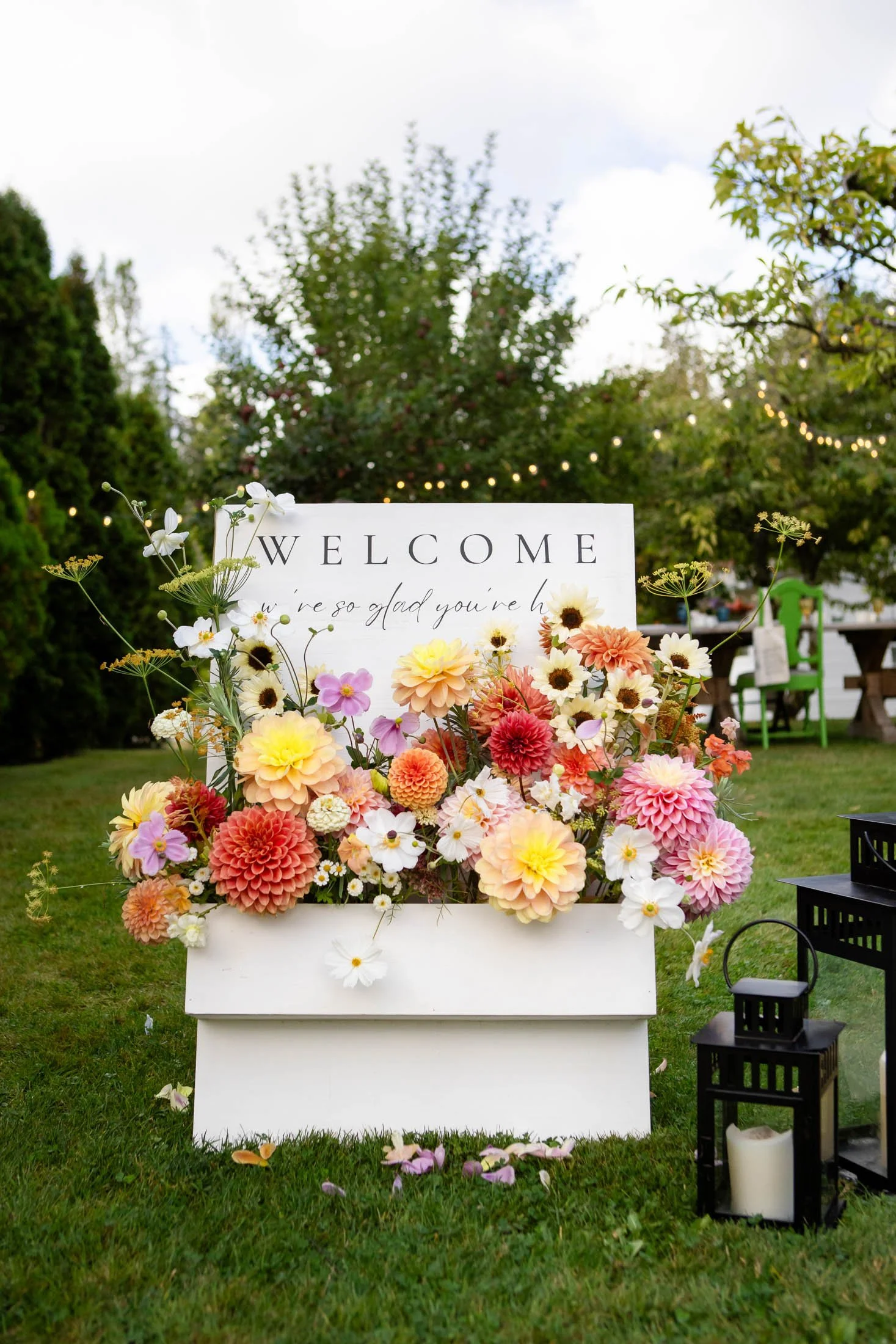 Flower arrangement with a sign that says "WELCOME" and a message underneath, placed outdoors with grass, trees, and string lights in the background. Candles and lanterns are on the ground nearby.
