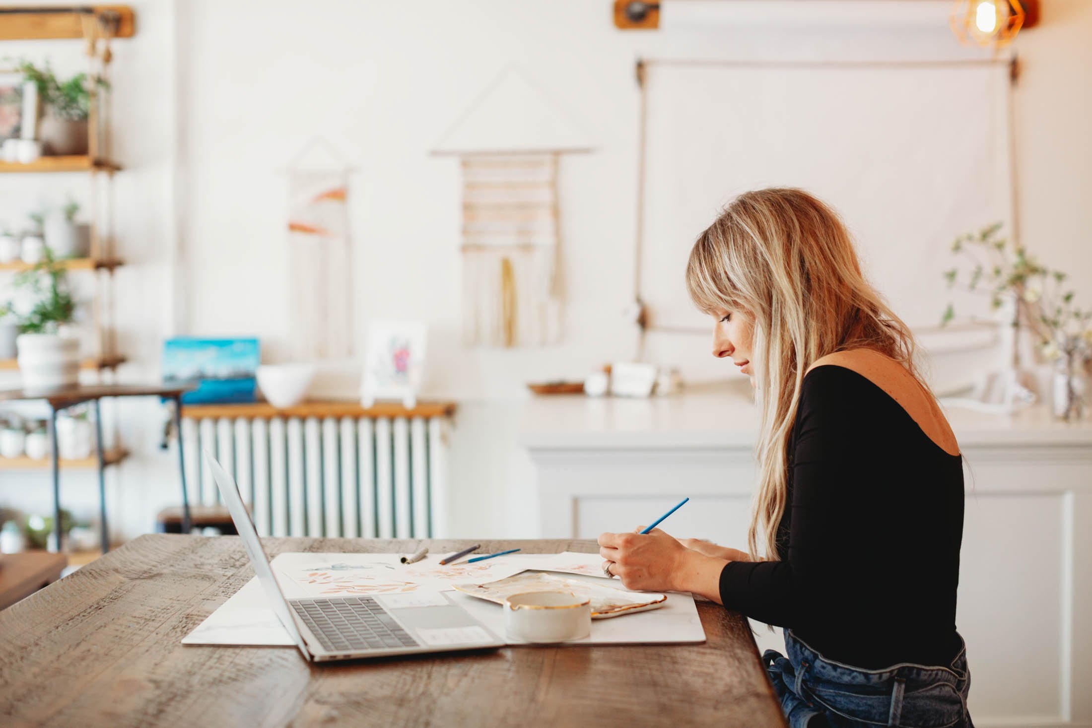 Woman with long, blonde hair sitting at a wooden table, working on art projects with paints and brushes, in a bright, cozy room with shelves, plants, and decorative wall hangings.