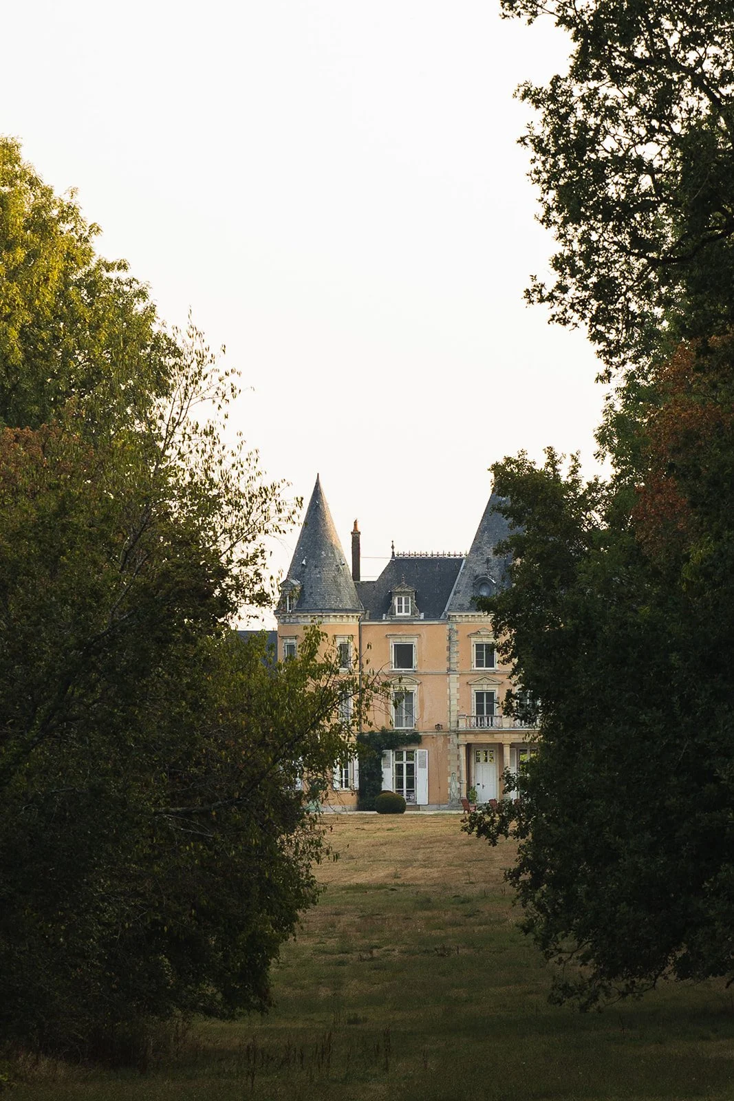 A castle-like house with pointed turrets, surrounded by trees, viewed through a natural frame of foliage, with a clear sky in the background.