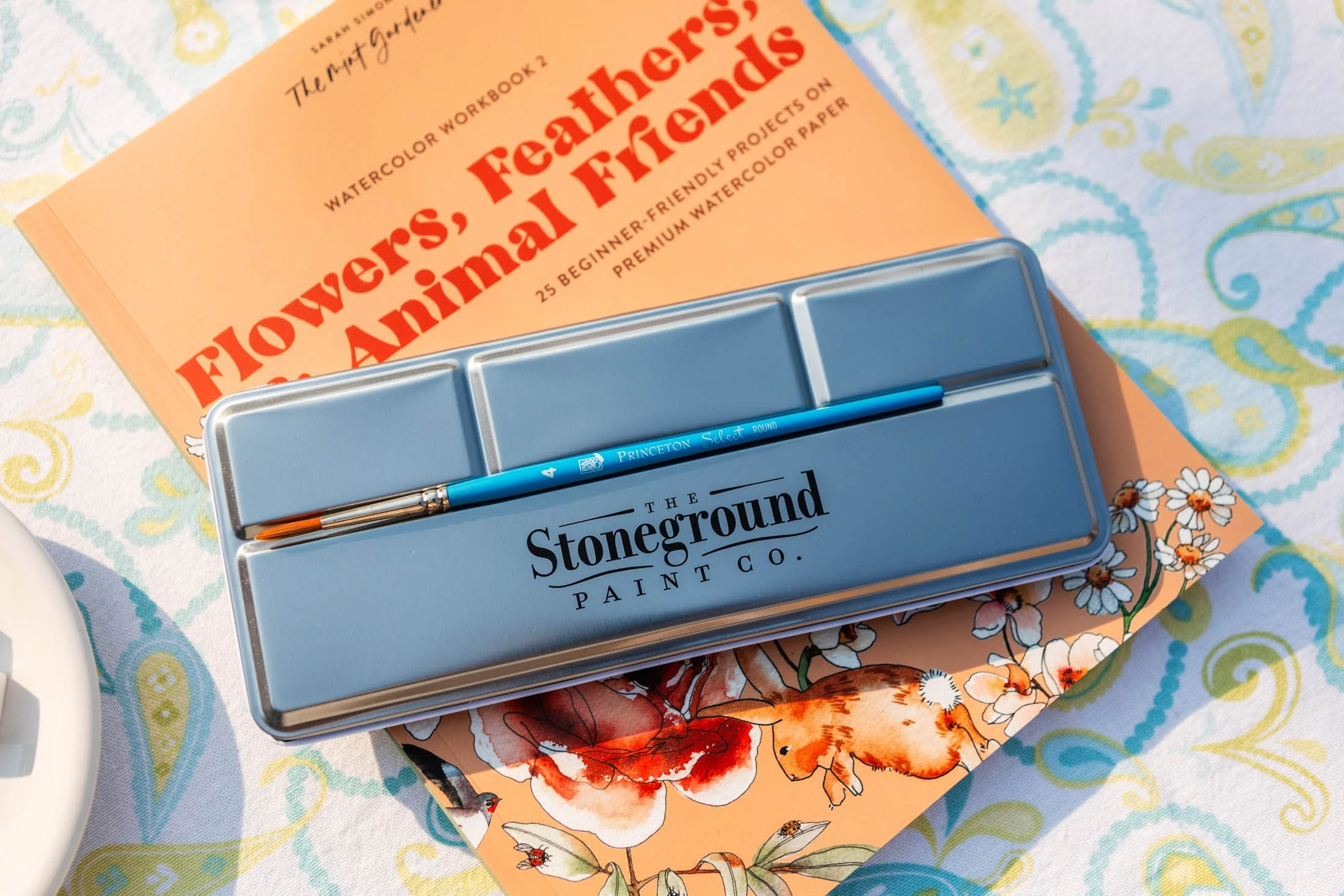 A water color painting book titled 'Flowers, Feathers, and Fairies' by Sarah Bickerdike, a paintbrush, and a light blue box labeled 'Stoneground Paint Co.' are placed on a table covered with a colorful patterned cloth.