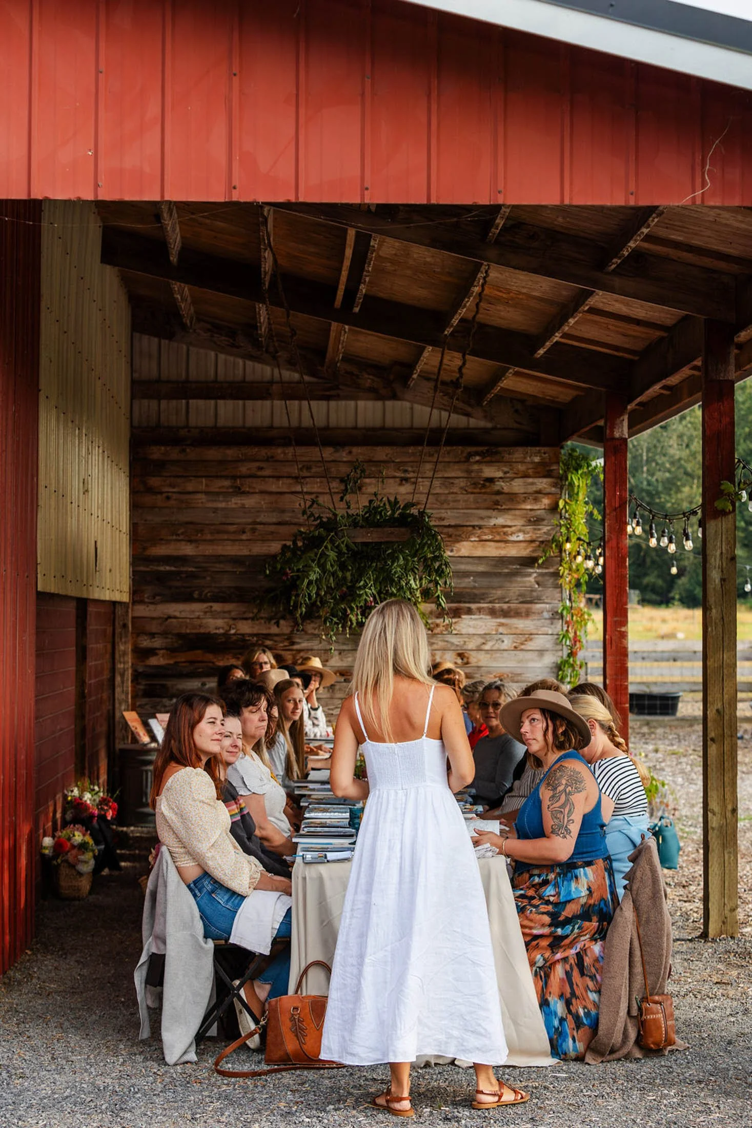 A group of people sitting at a long table under a rustic wooden and metal pavilion, listening to a woman in a white dress speaking to the crowd. The setting is outdoors with greenery in the background, and there are string lights hanging from the pavilion.
