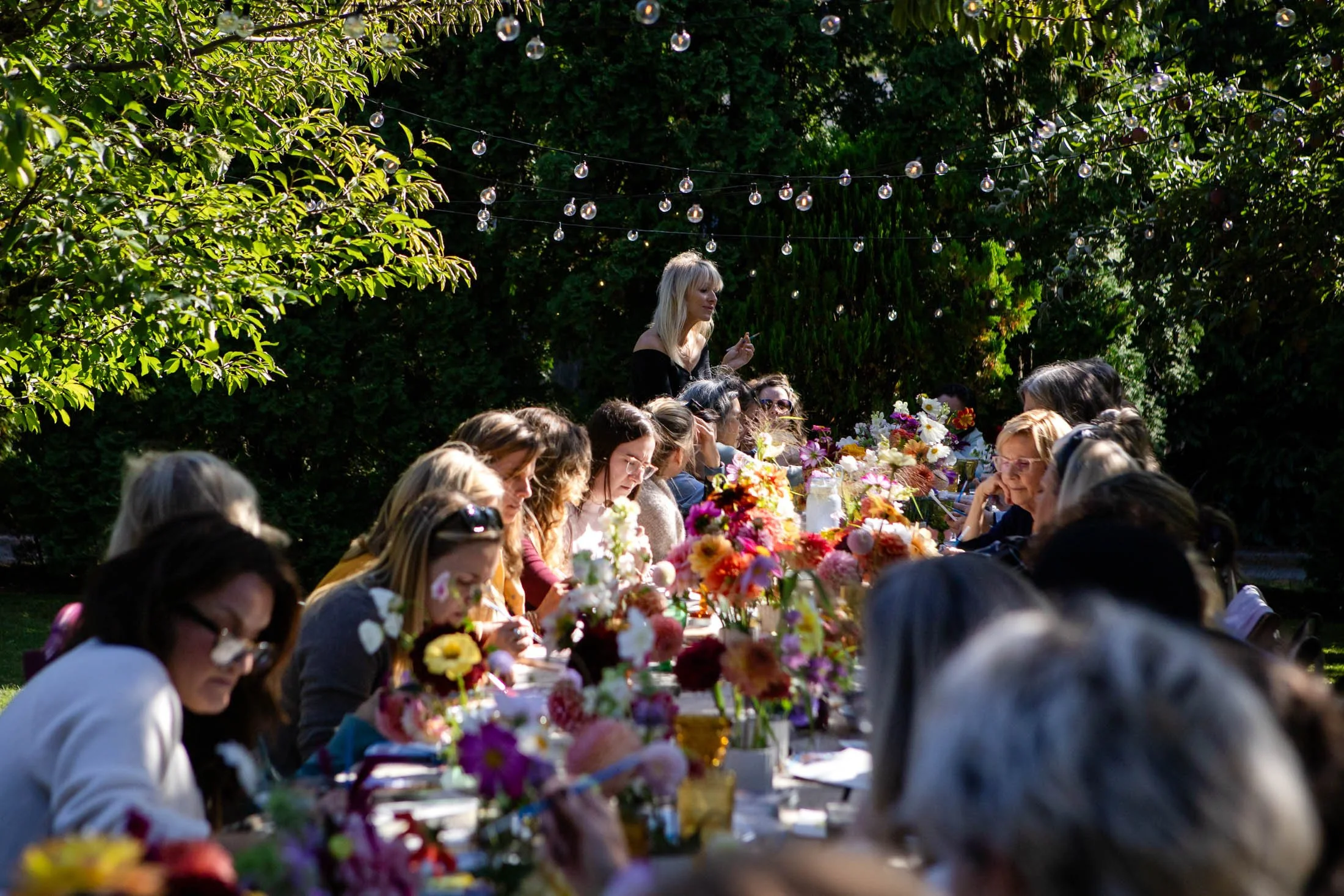 People seated at an outdoor event table decorated with colorful flowers, with a woman standing and speaking, surrounded by lush green trees and string lights overhead.