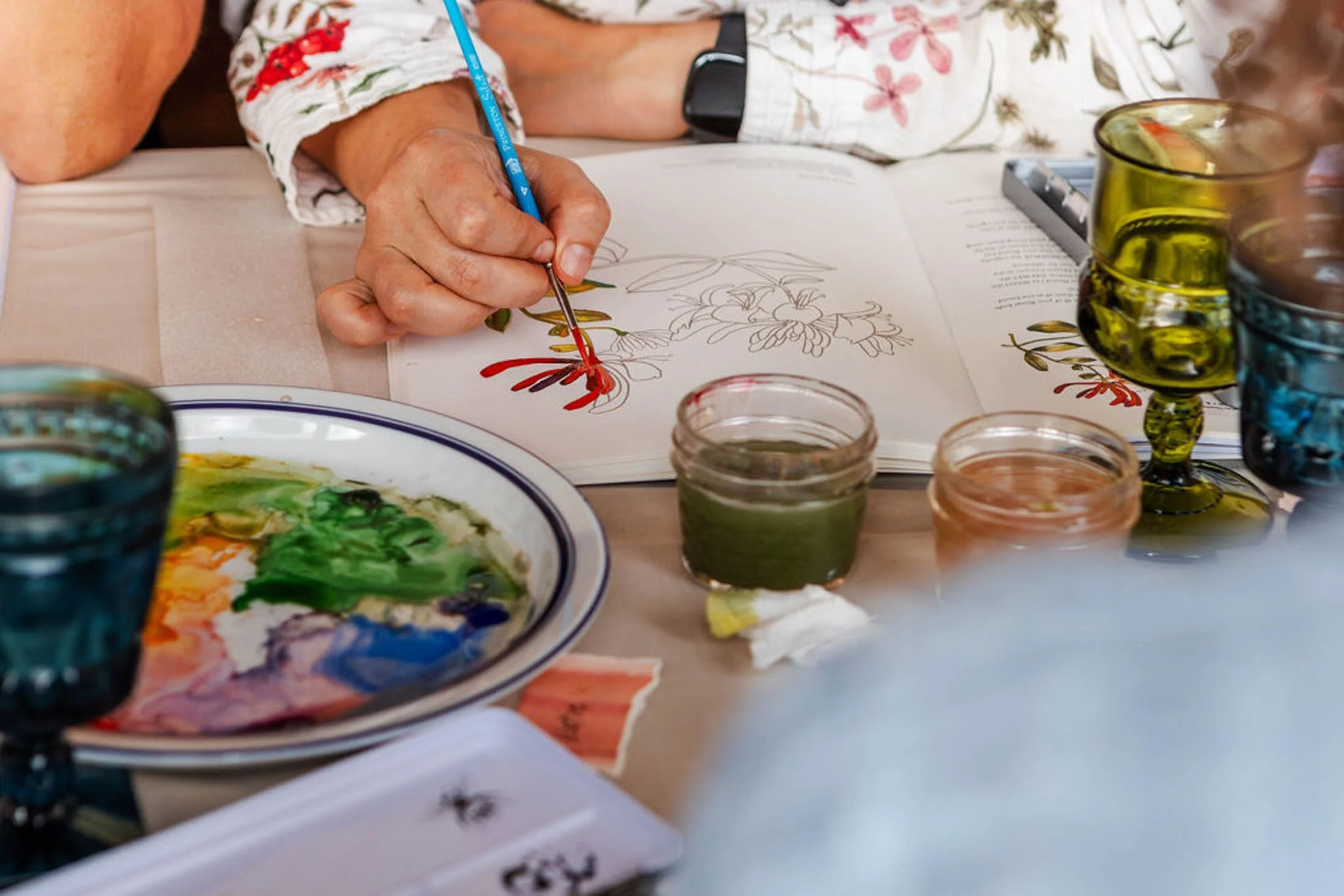 Person painting botanical illustrations with watercolors, surrounded by glass jars of paint and paintbrushes.
