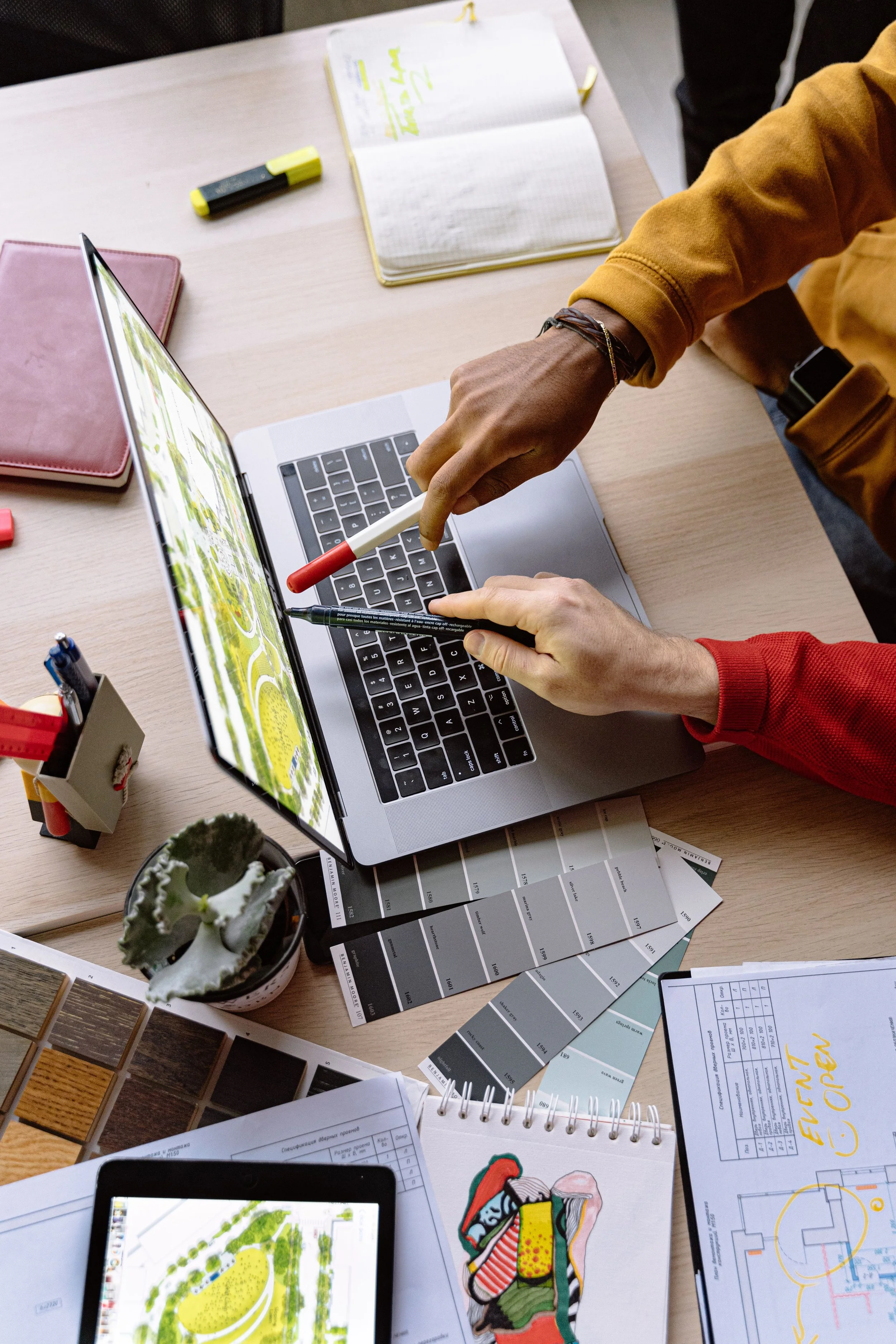 Hands at a creative desk with notebooks, color swatches and a tablet.