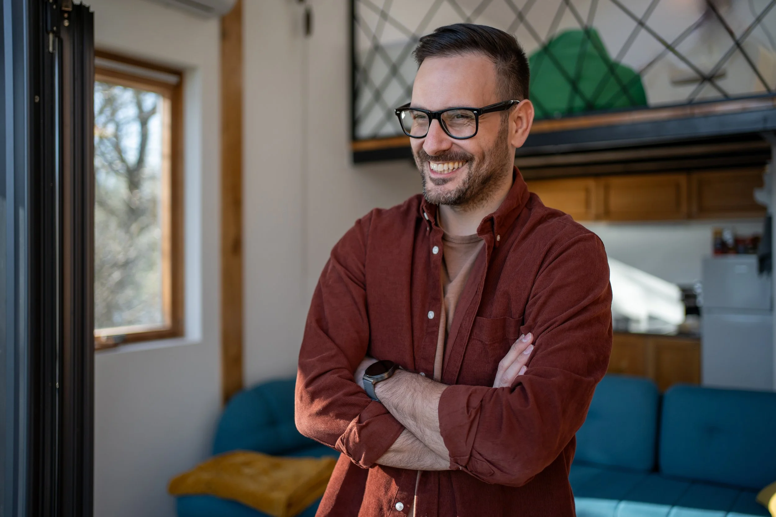 A smiling man with glasses standing near a window.