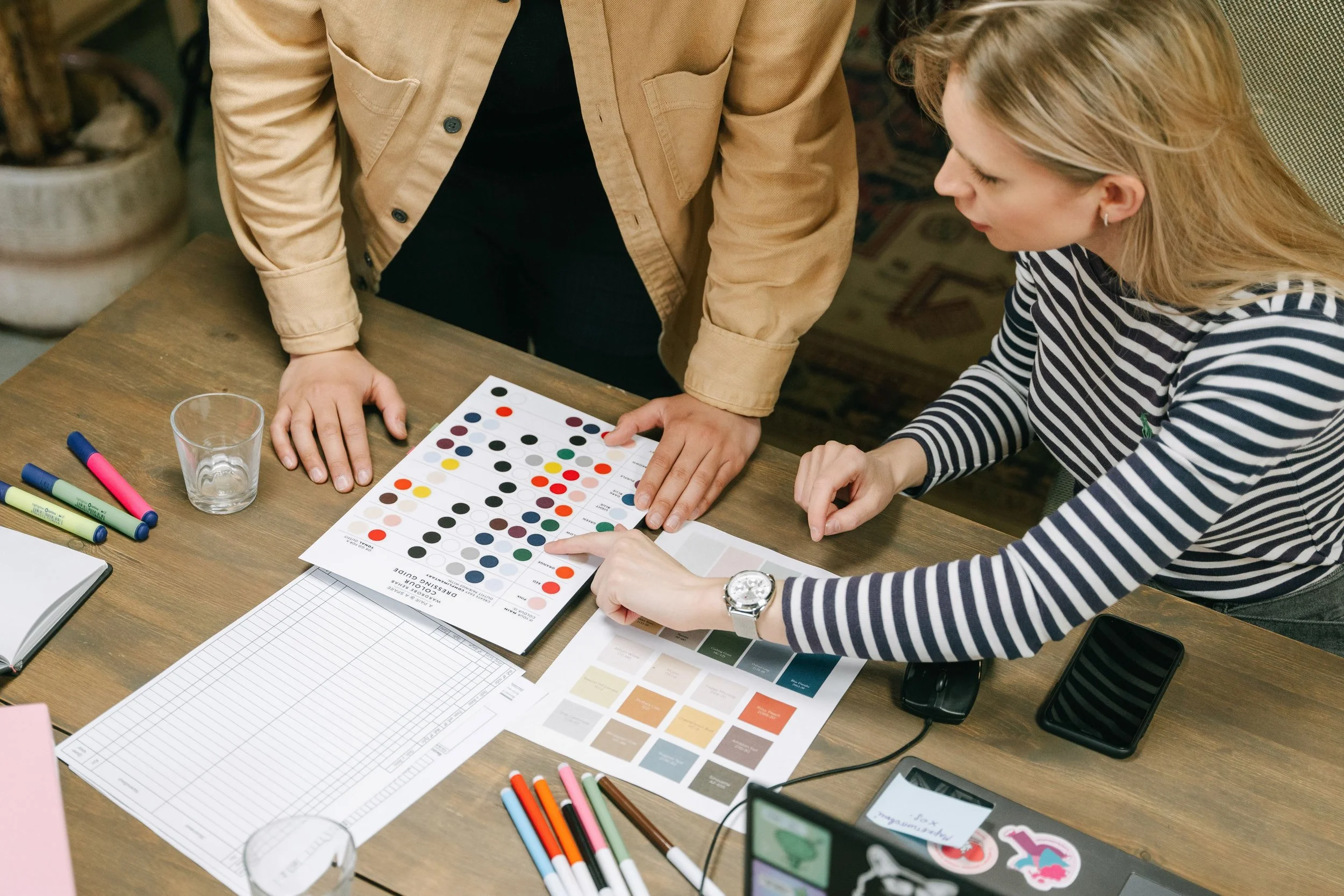 Two people are examining a color guide chart on a wooden table with markers, a notepad, and a glass around them. One person is pointing at the chart, and the other is looking at it.