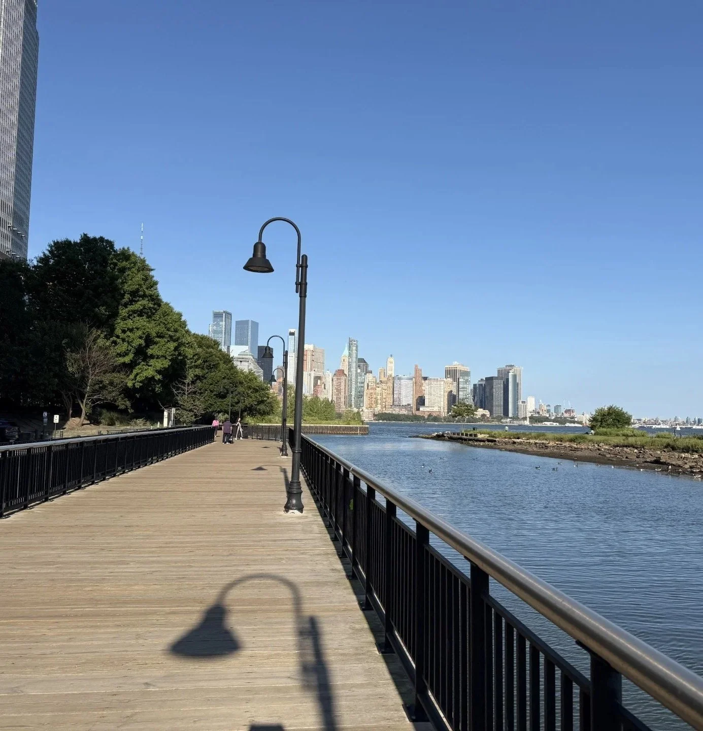 A wooden walkway beside a body of water with city skyline in the background, trees on the left, lampposts on the walkway, and two people walking in the distance on a bright, clear day.