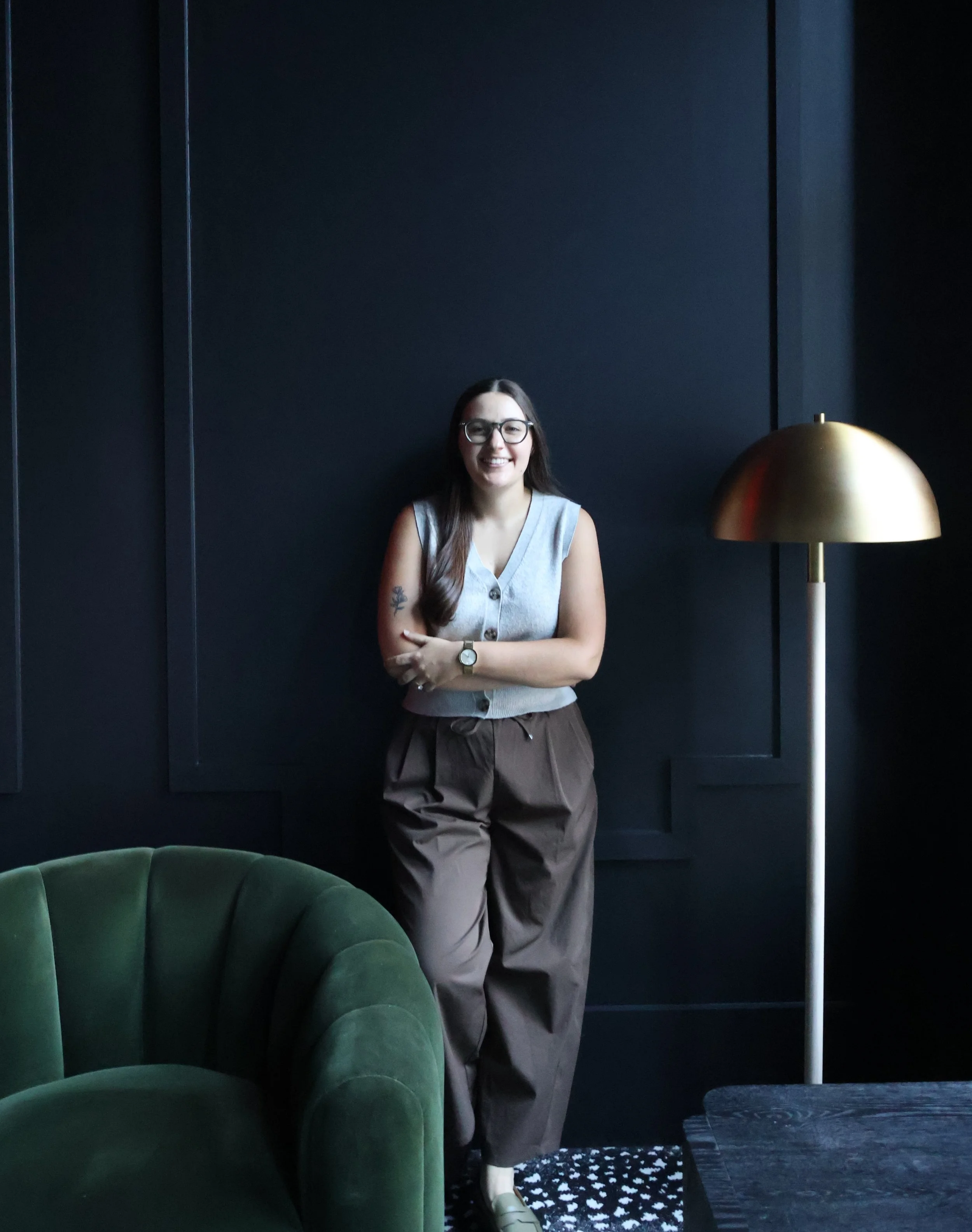 A woman standing indoors, smiling, wearing glasses, a sleeveless gray vest, brown pants, and beige shoes, beside a gold-colored floor lamp and green velvet seating.