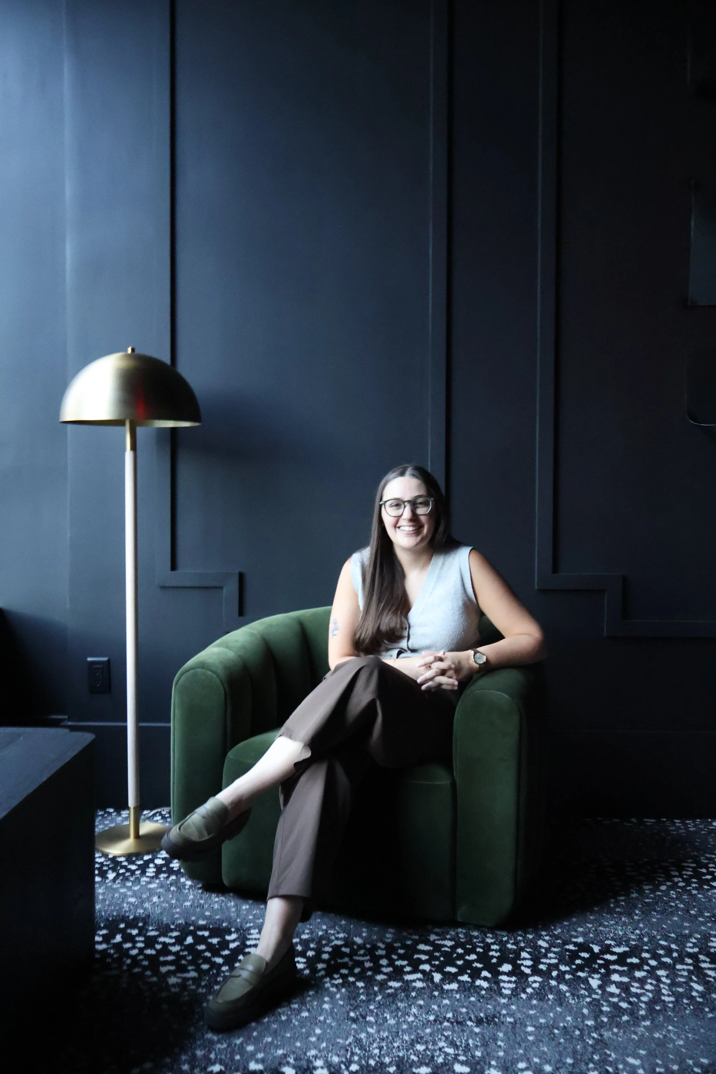 A woman with glasses is sitting on a green armchair in a dark, modern room, smiling. There is a gold-colored floor lamp beside her, and a dark wall with paneling behind her.