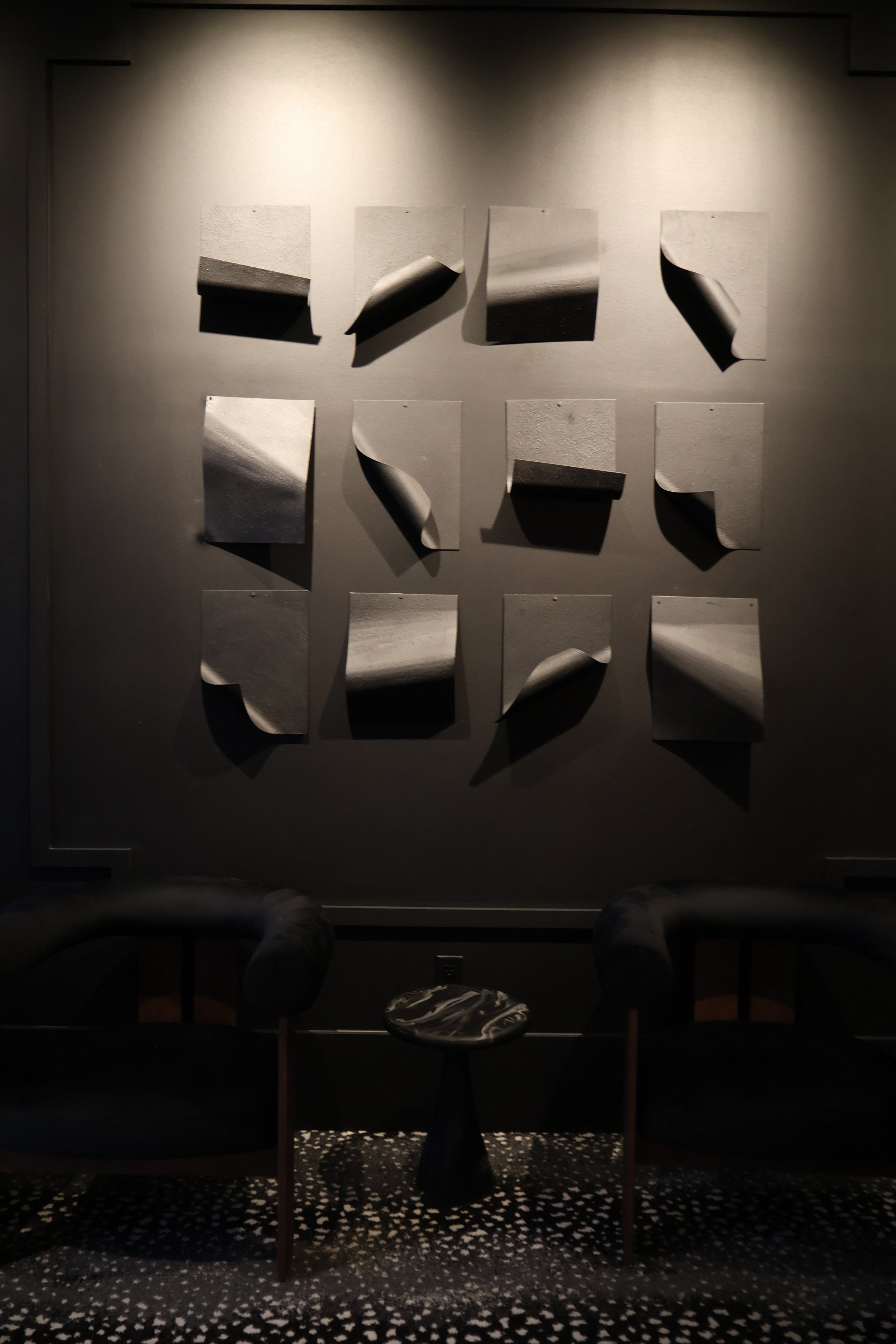 Photo taken in a dark room with a wall displaying nine black and white abstract paper art pieces. The art pieces have various shapes and textures, with some curling or bending. Two chairs are visible at the bottom of the image, facing the wall.