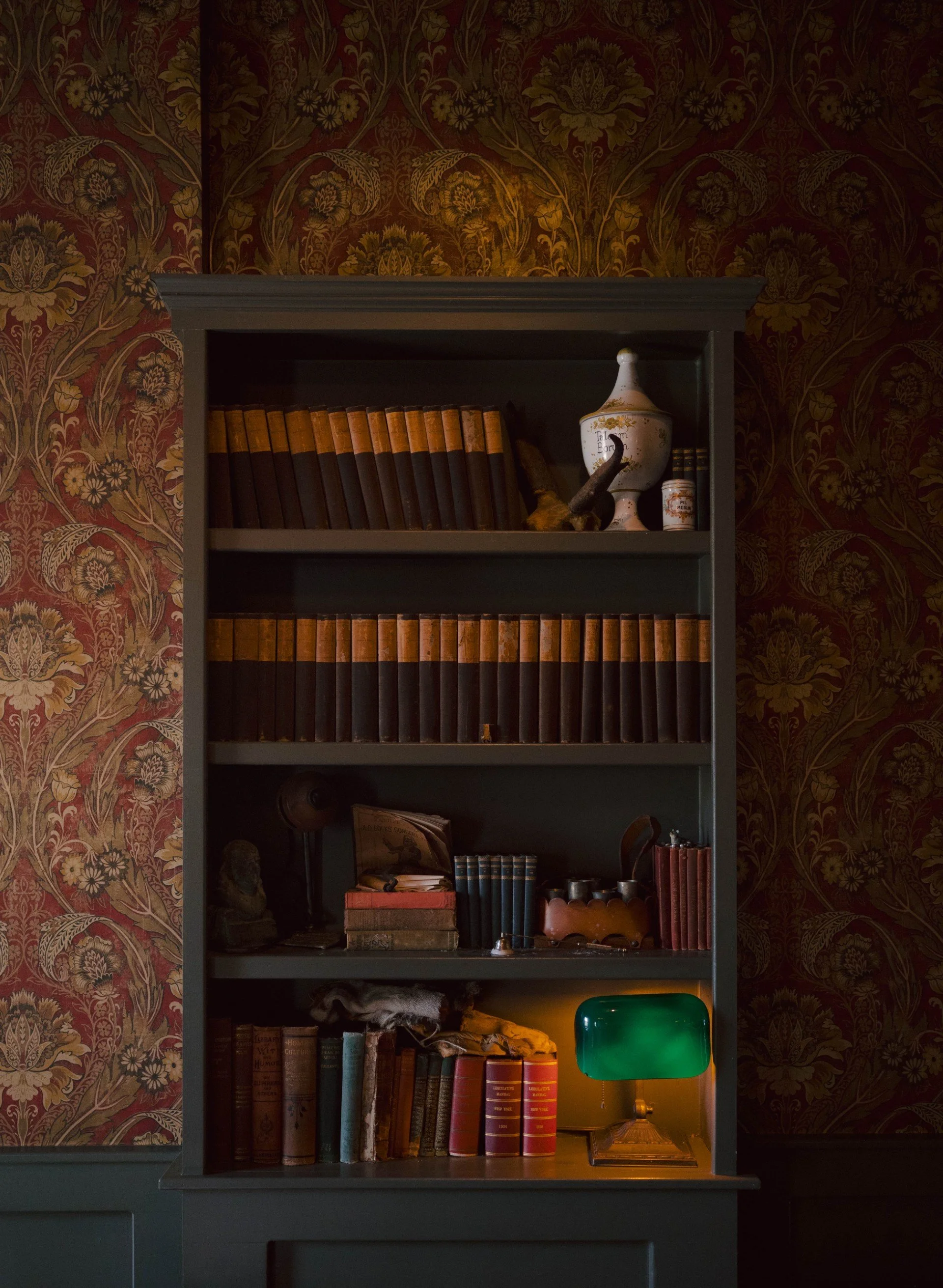 Bookshelf filled with antique books, a decorative urn, and a green desk lamp, against floral patterned wallpaper in a dimly lit room.