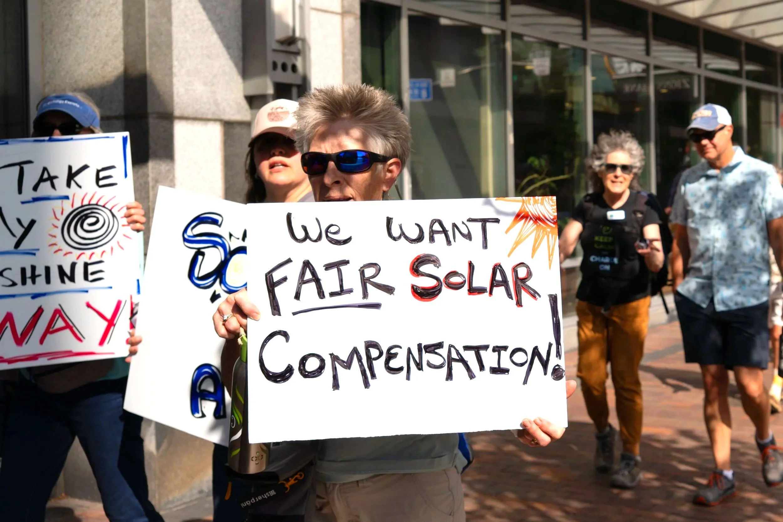 eoman with short hair and sunglasses holding a sign that says "We want Fair Solar Compensation"