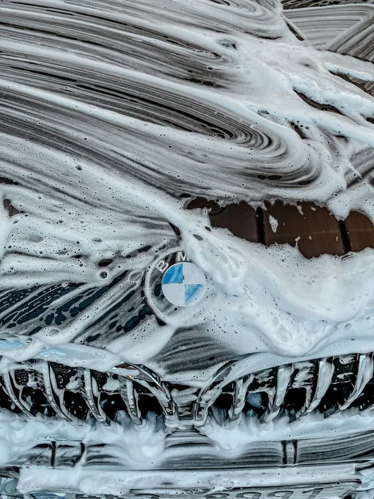 A BMW car front grille covered in soap suds during washing, with the BMW logo visible.