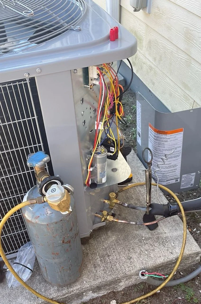 An outdoor HVAC unit with open service panel, exposing electrical wiring and a refrigerant cylinder connected to the system.