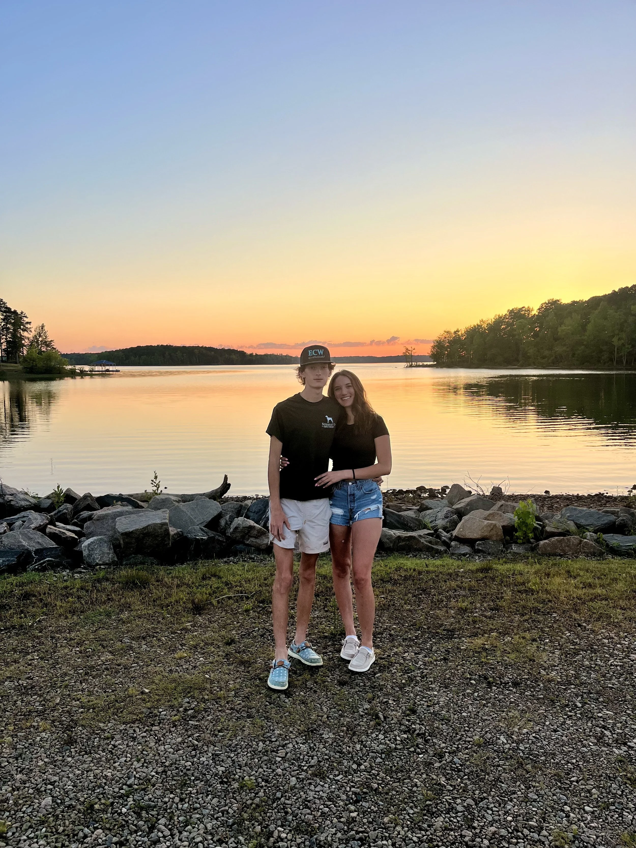A young man and woman standing on rocks by a lake during sunset, smiling and embracing each other.