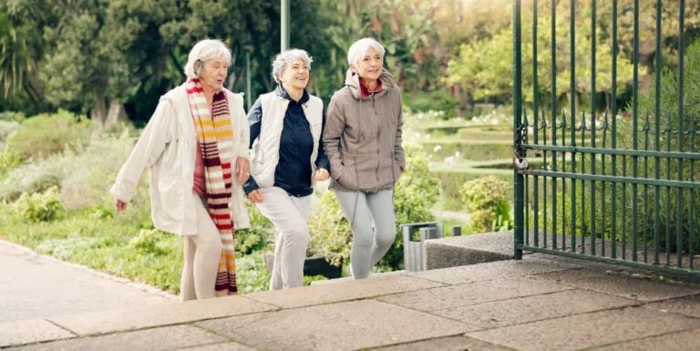 Three elderly women wearing coats and scarves walking outdoors near a metal gate in a park or garden during daytime.