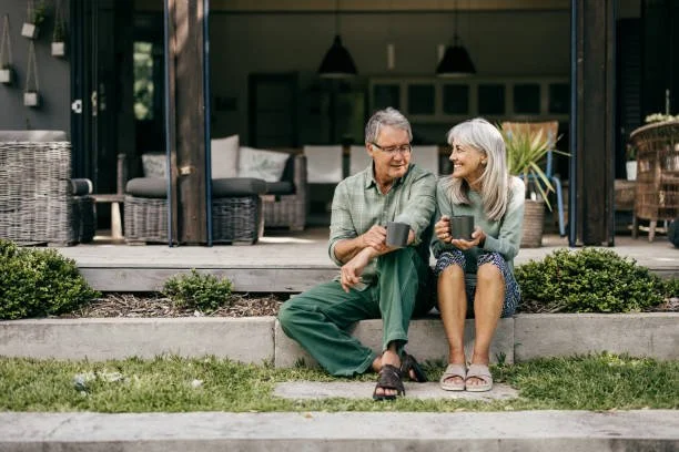 An elderly couple sitting on the steps outside their house, holding coffee mugs and smiling at each other.