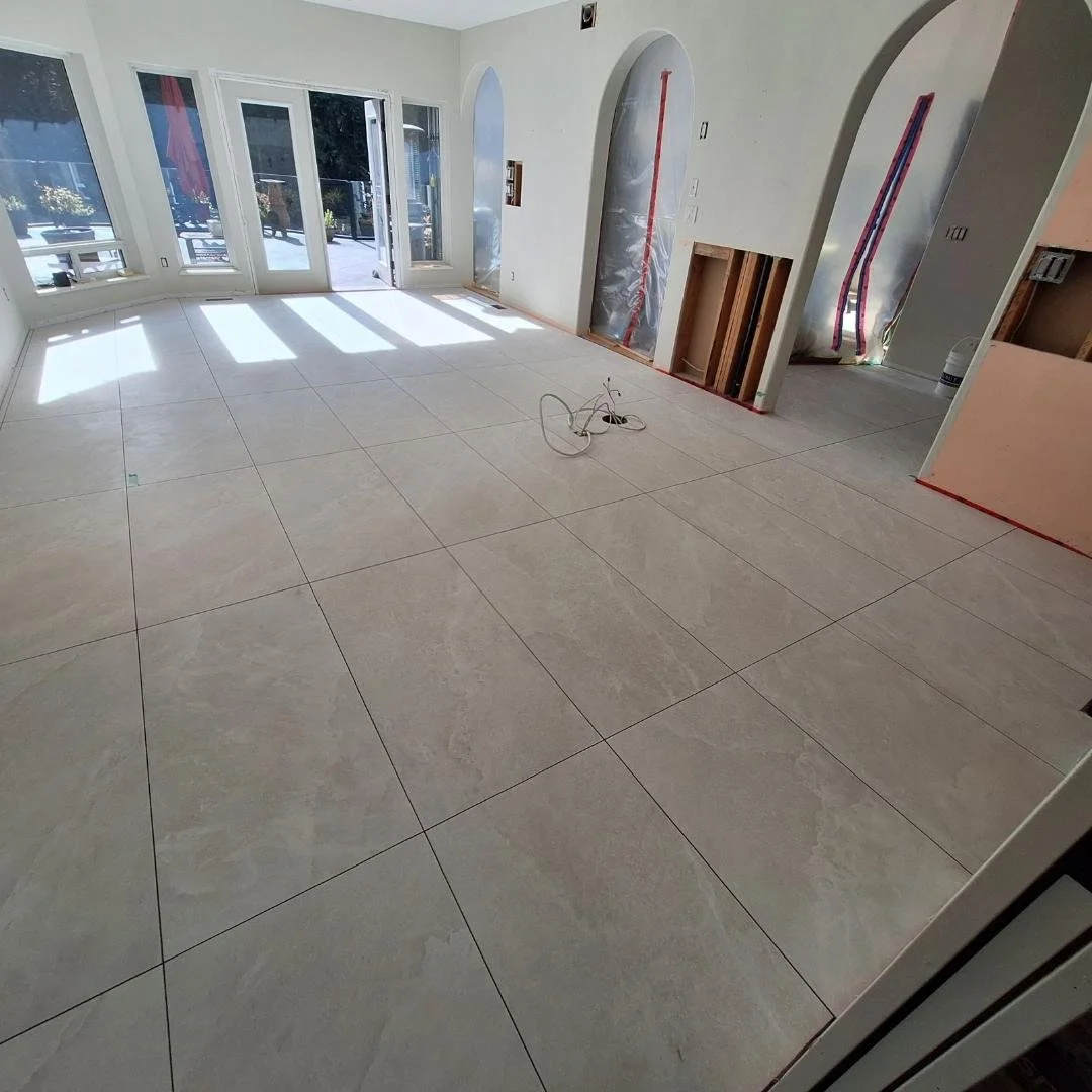 An interior room under construction with large light-colored tile flooring, white walls, and large windows with sunlight streaming in. Some electrical wiring and construction materials are visible.
