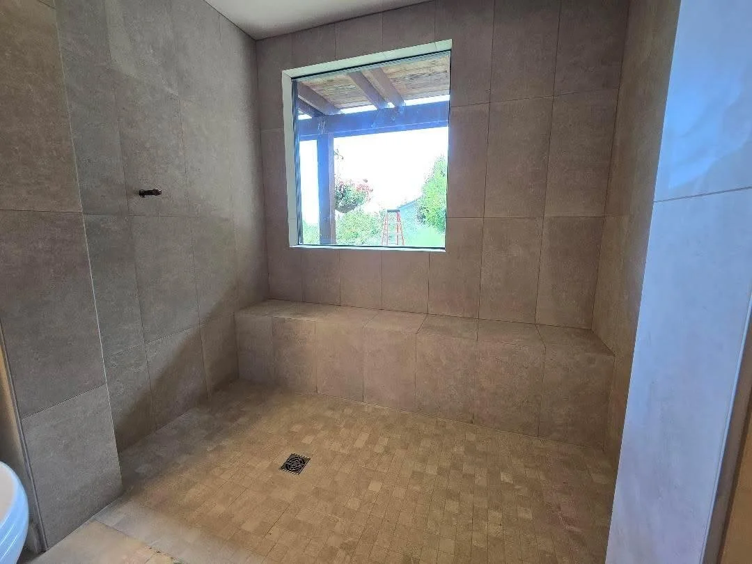 A partially finished tiled shower with beige tiles, a window, and a built-in bench, with a view of an outdoor area with trees and a ladder outside.