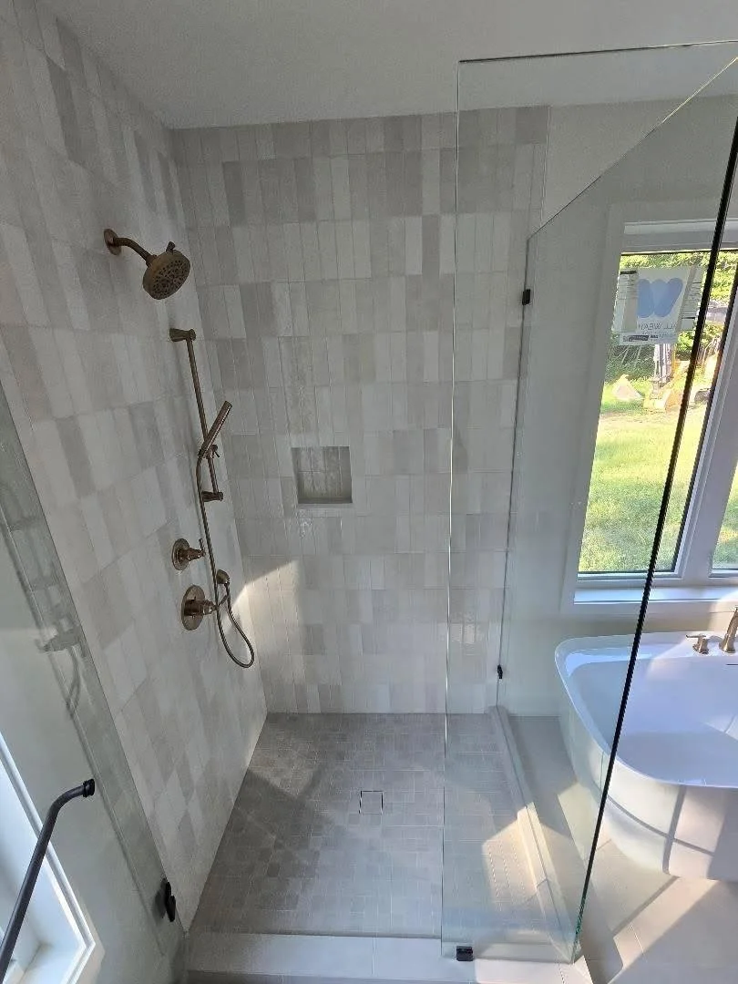 Shower with rain shower head, handheld shower, and gray tile walls separated by a glass partition, alongside a window and a sink