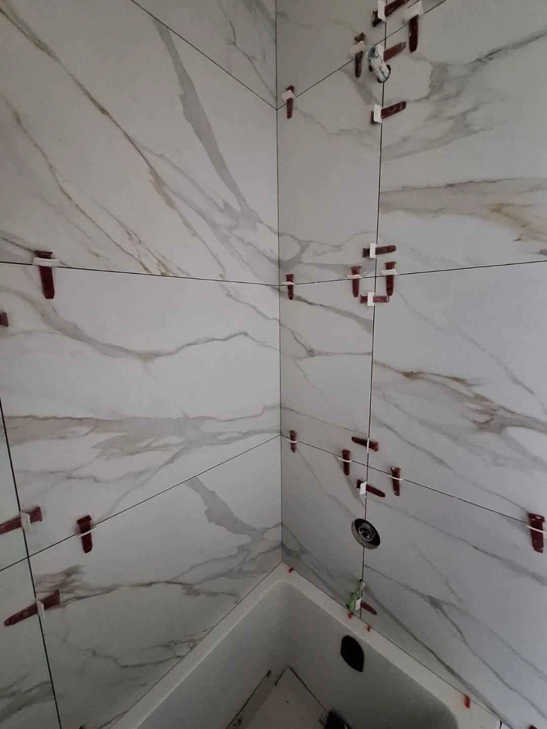 Corner of a bathroom with marble-like wall tiles, showing tile spacers and a drain at the bottom, indicating ongoing tiling work.