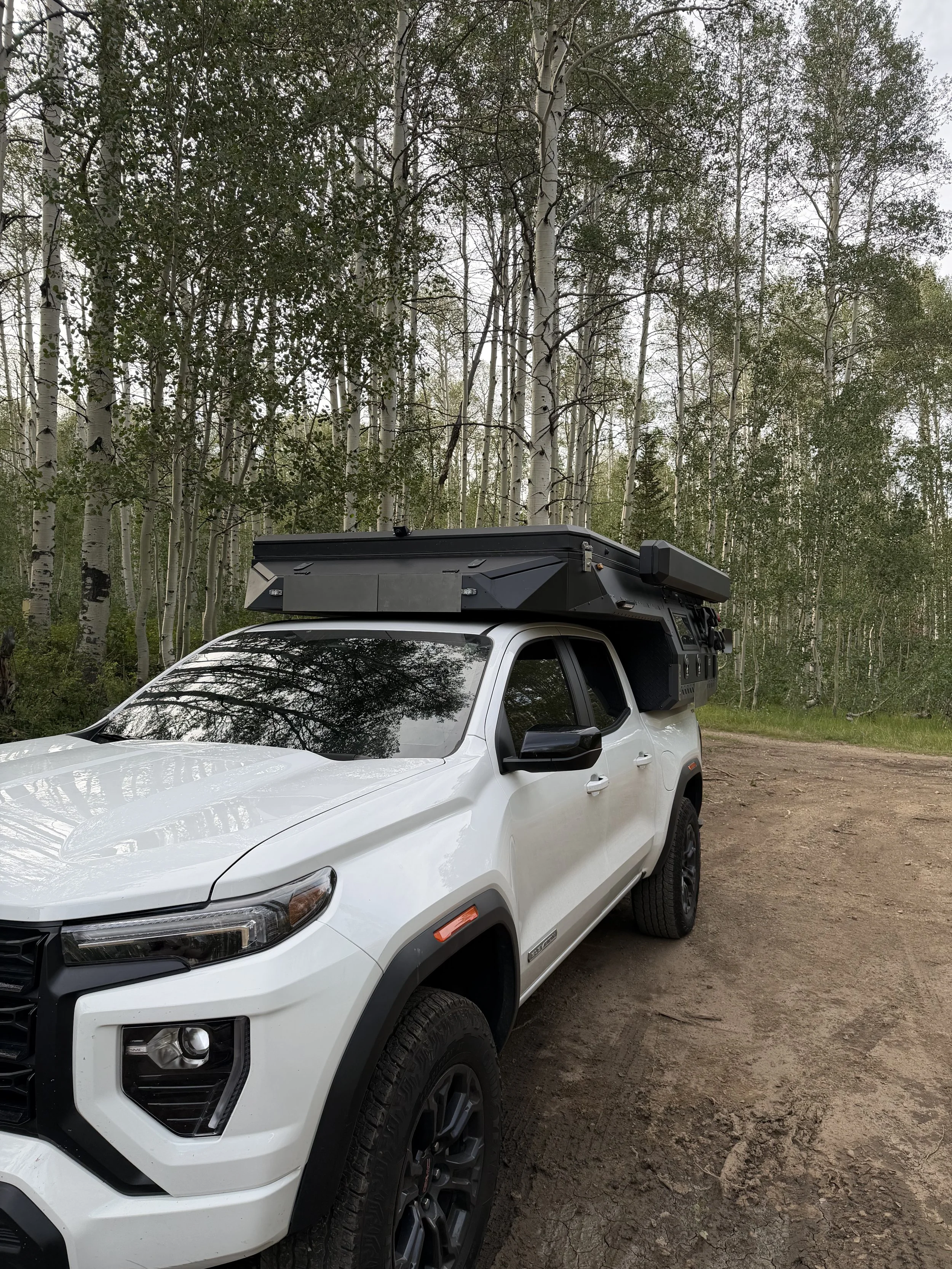 White pickup truck equipped with mounted electronic or communication equipment parked on a dirt road in front of a forest of tall, white-barked trees.