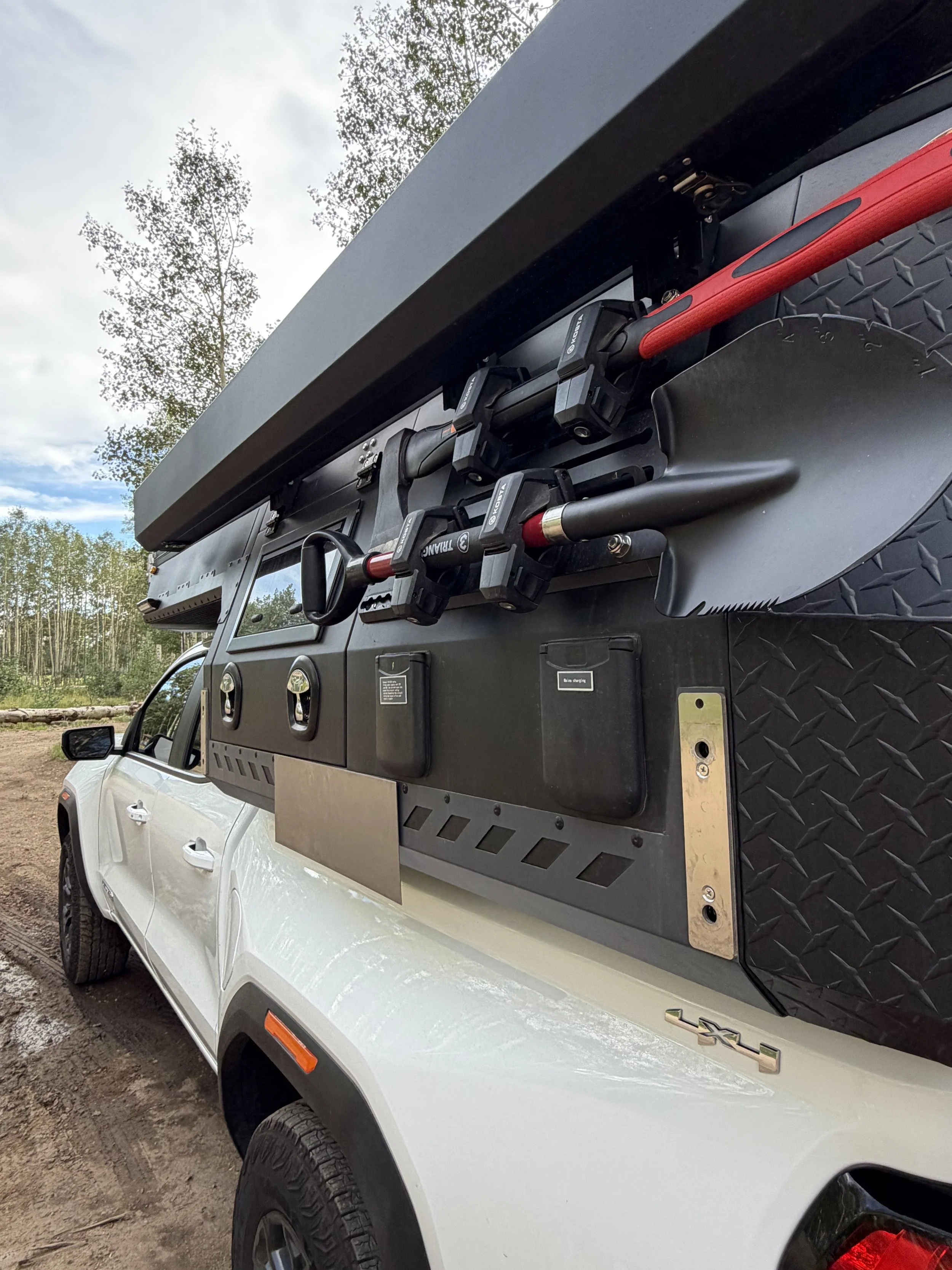Close-up of a white truck with specialized equipment and tools mounted on the side, including cables, a black box, and a spare wheel, parked on a dirt road with trees and a cloudy sky in the background.