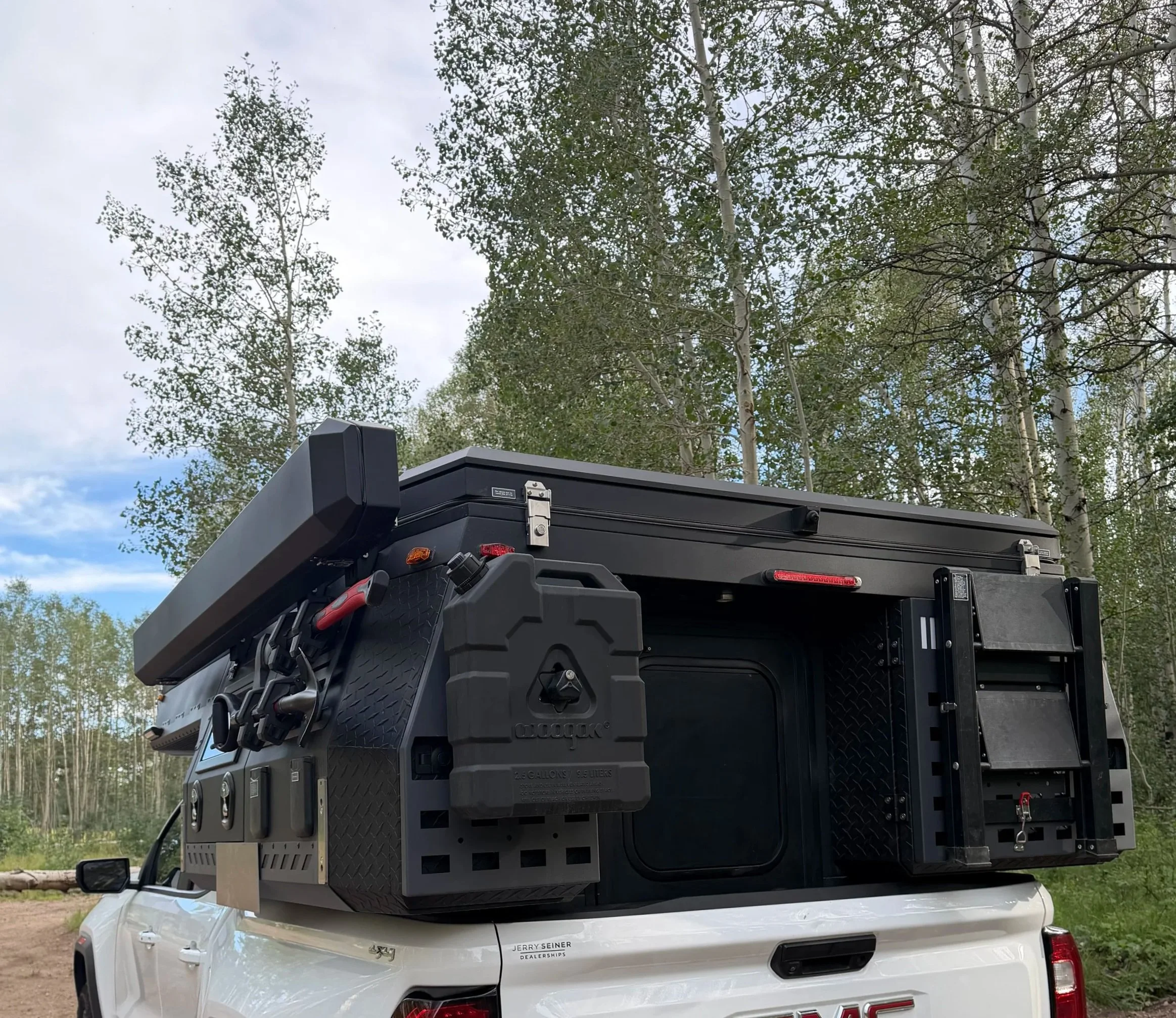 The image shows the back of a white GMC truck with a large black equipment box mounted on the bed. The box has various compartments and equipment attached, including hoses and connectors. The truck is outdoors in a wooded area with tall trees and a cloudy sky in the background.