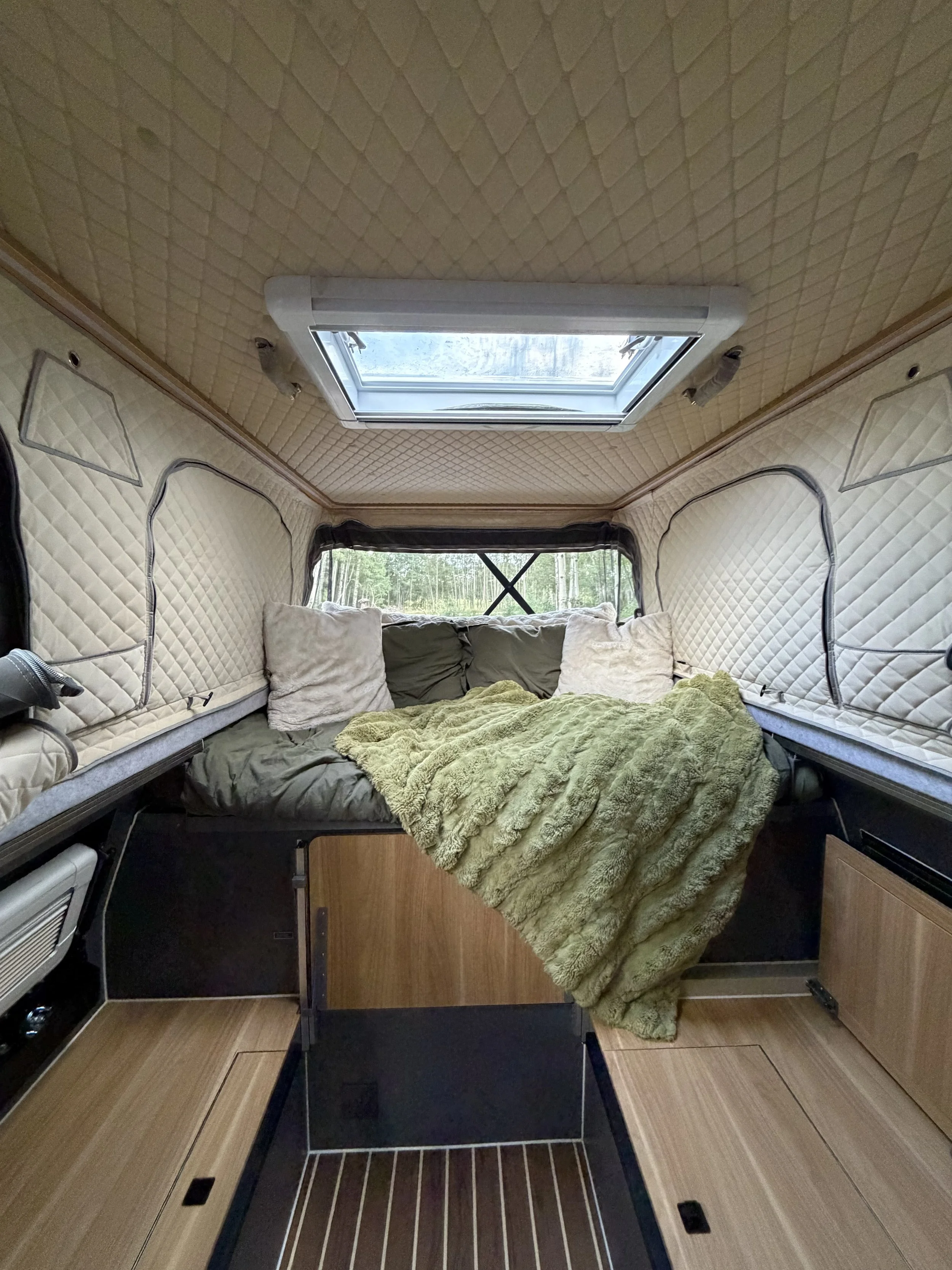 Interior of a camper or RV with beige quilted walls, a skylight in the ceiling, a bed with pillows and a green blanket, and a small wooden table.
