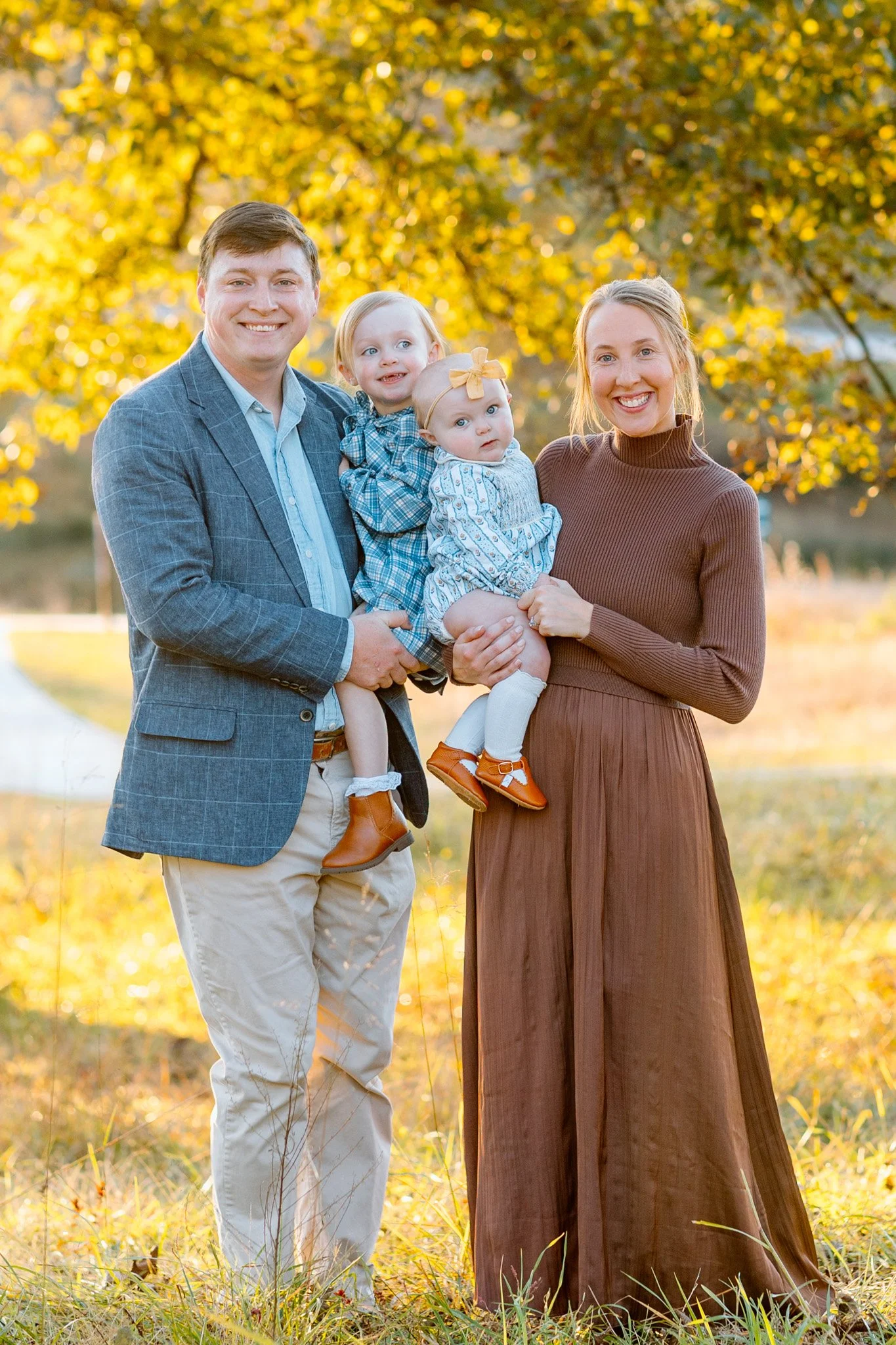 A family of four standing outdoors in fall, with yellow leaves in the background. The father is on the left in a blue plaid blazer and khakis, holding two young daughters. The mother is on the right in a brown dress, holding one of the daughters. All are smiling and looking at the camera.