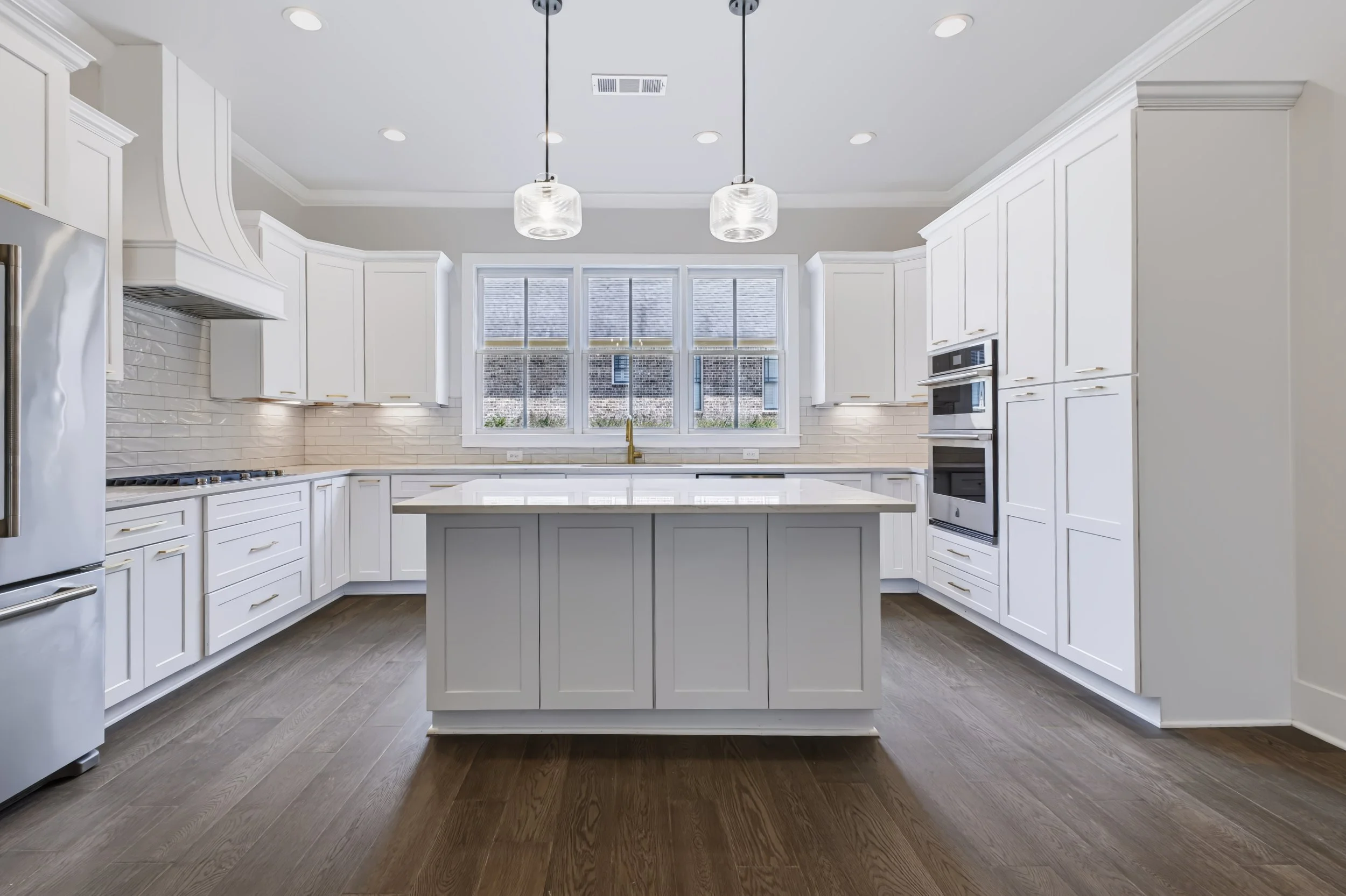Modern white kitchen with large center island, white cabinets, stainless steel refrigerator, built-in oven, gas cooktop, and large windows.