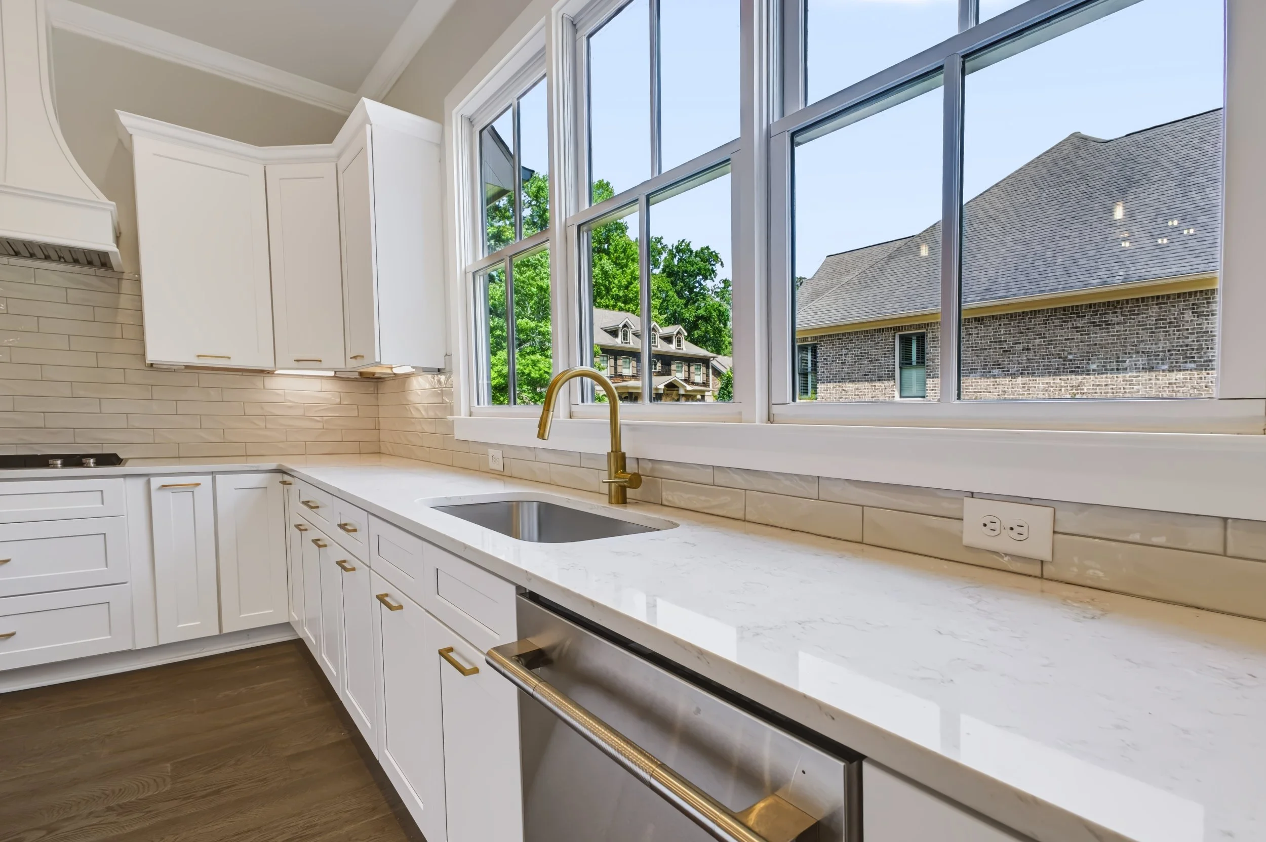 Bright kitchen with large window, white cabinets, beige backsplash, marble countertop, gold fixtures, and view of neighboring houses and green trees.