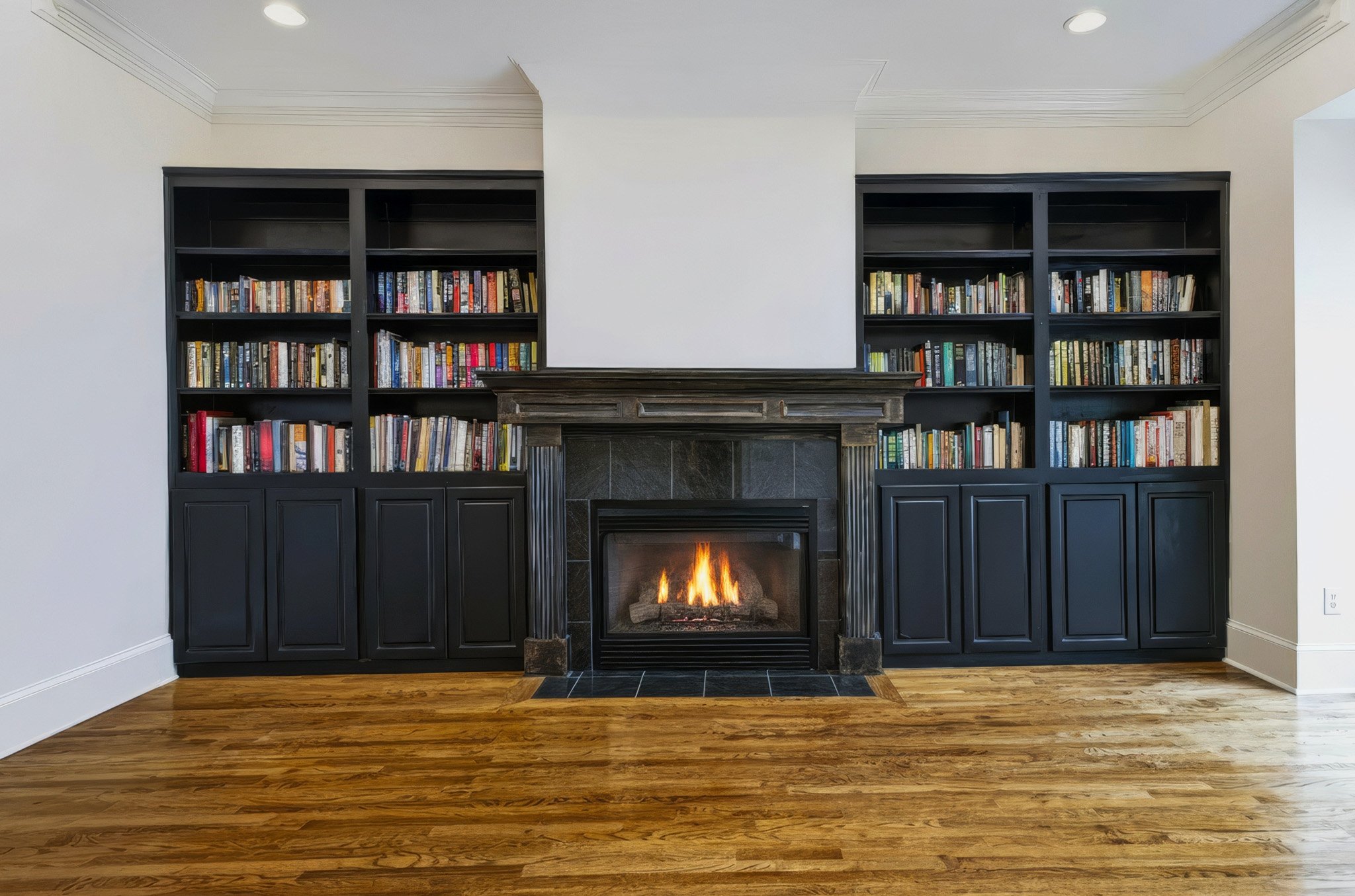 A living room with a fireplace flanked by black bookshelves filled with books, wood flooring, and white walls with crown molding.