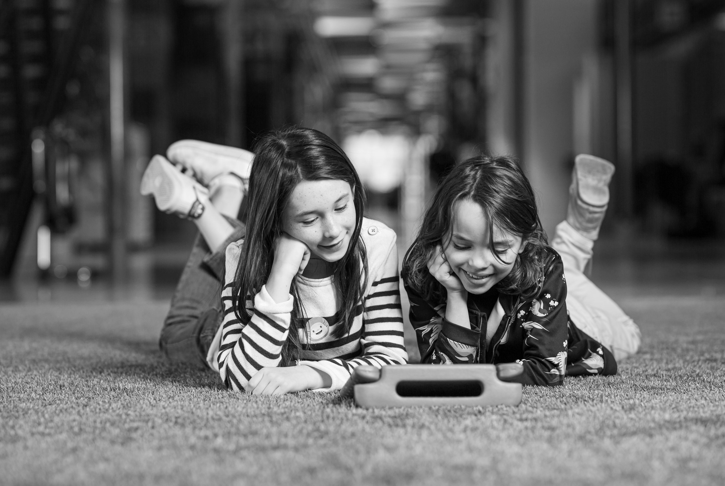 Two young girls lying on their stomachs on a carpeted floor, looking at a tablet and smiling, with one girl resting her chin on her hand and the other gently touching the tablet.