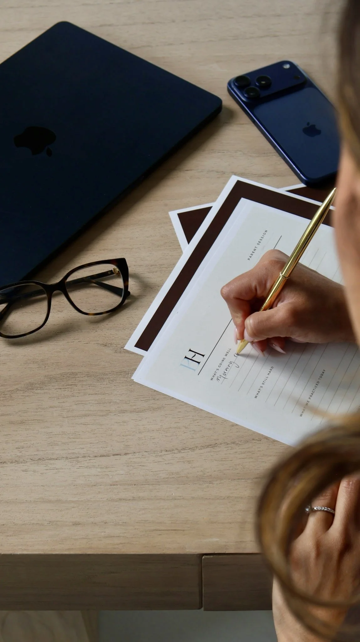 A person writing on a form labeled "Parent Session" at a desk with a closed laptop, a smartphone, glasses, and photographs.