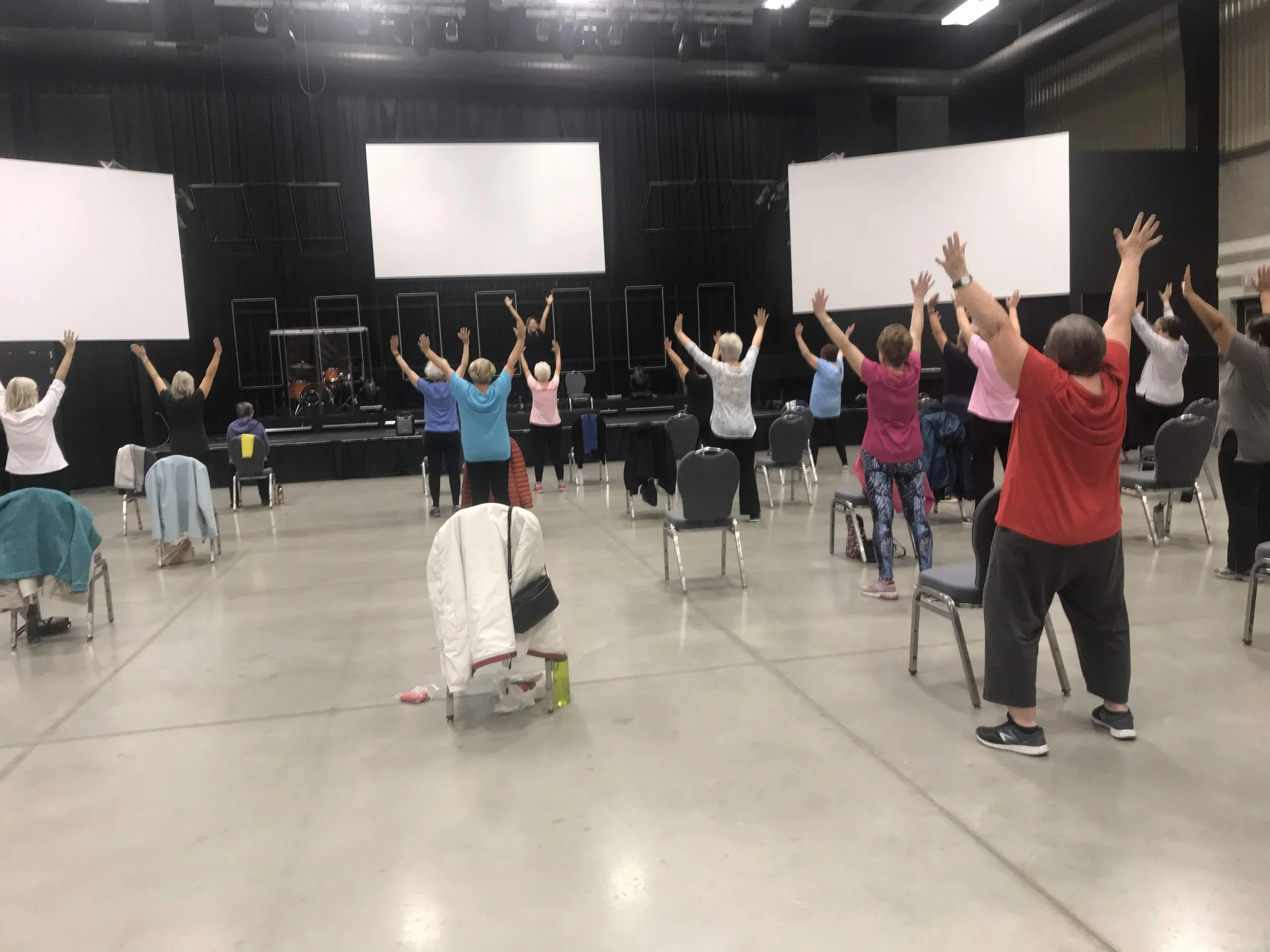 Group of people participating in a seated exercise or stretching class with arms raised, in a large room with black walls, large white screens, and chairs.