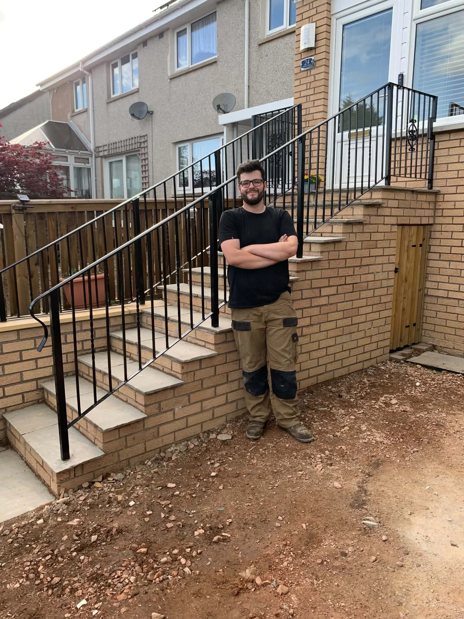 A smiling man with glasses and a beard standing outside on a construction site next to a brick staircase and new black metal railing, with a house in the background.