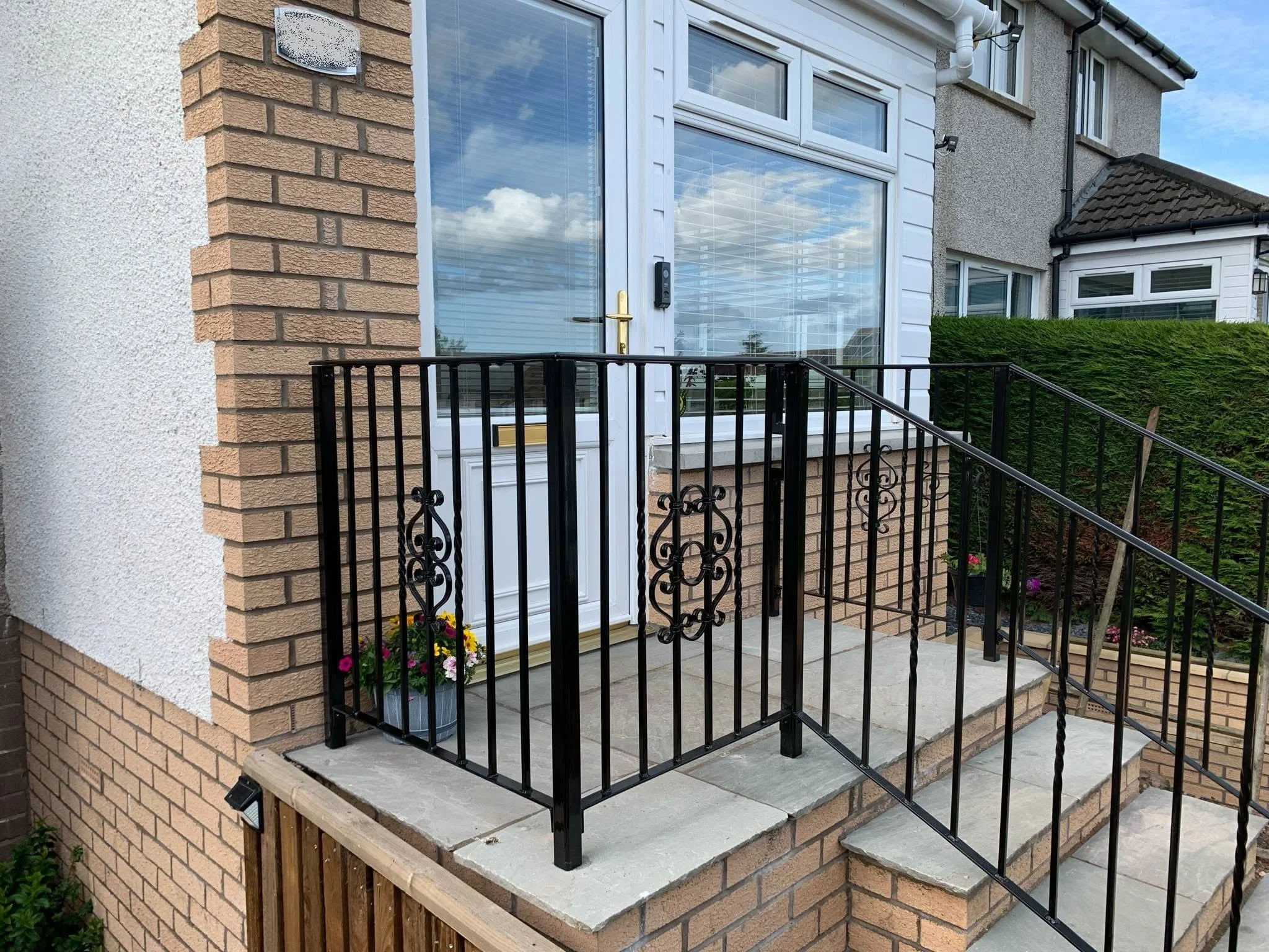 Front porch of a house with a glass door, brick walls, and black metal railing. There are flowers in a pot and a hedge nearby.