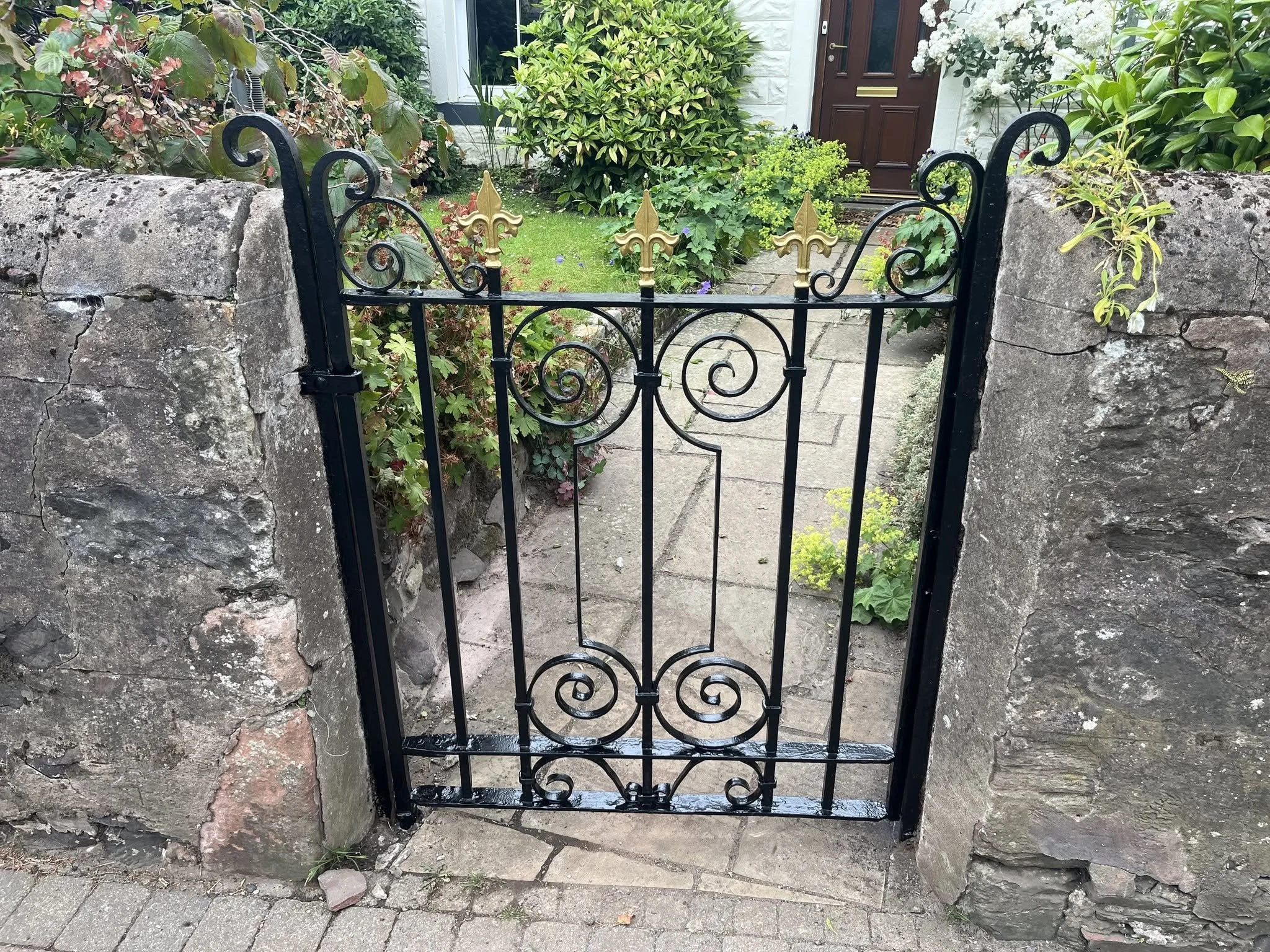 A wrought iron gate with gold accents set between stone pillars, leading to a garden with various green plants and a brick pathway.