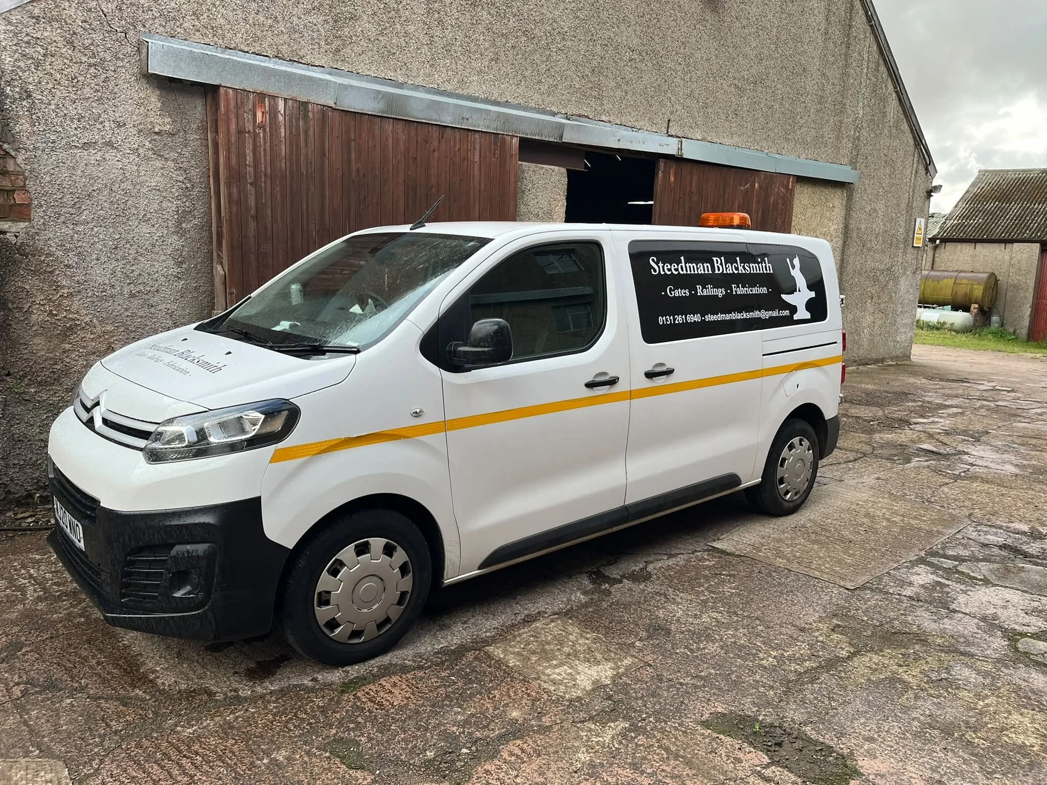 A white marketing van with orange and black markings parked outside a building with a textured concrete wall and a wooden sliding door. The van has the text 'Steedman Blacksmith' and services like gates, railings, and fabrication, along with a phone number and email. There is an orange warning light on top of the van.